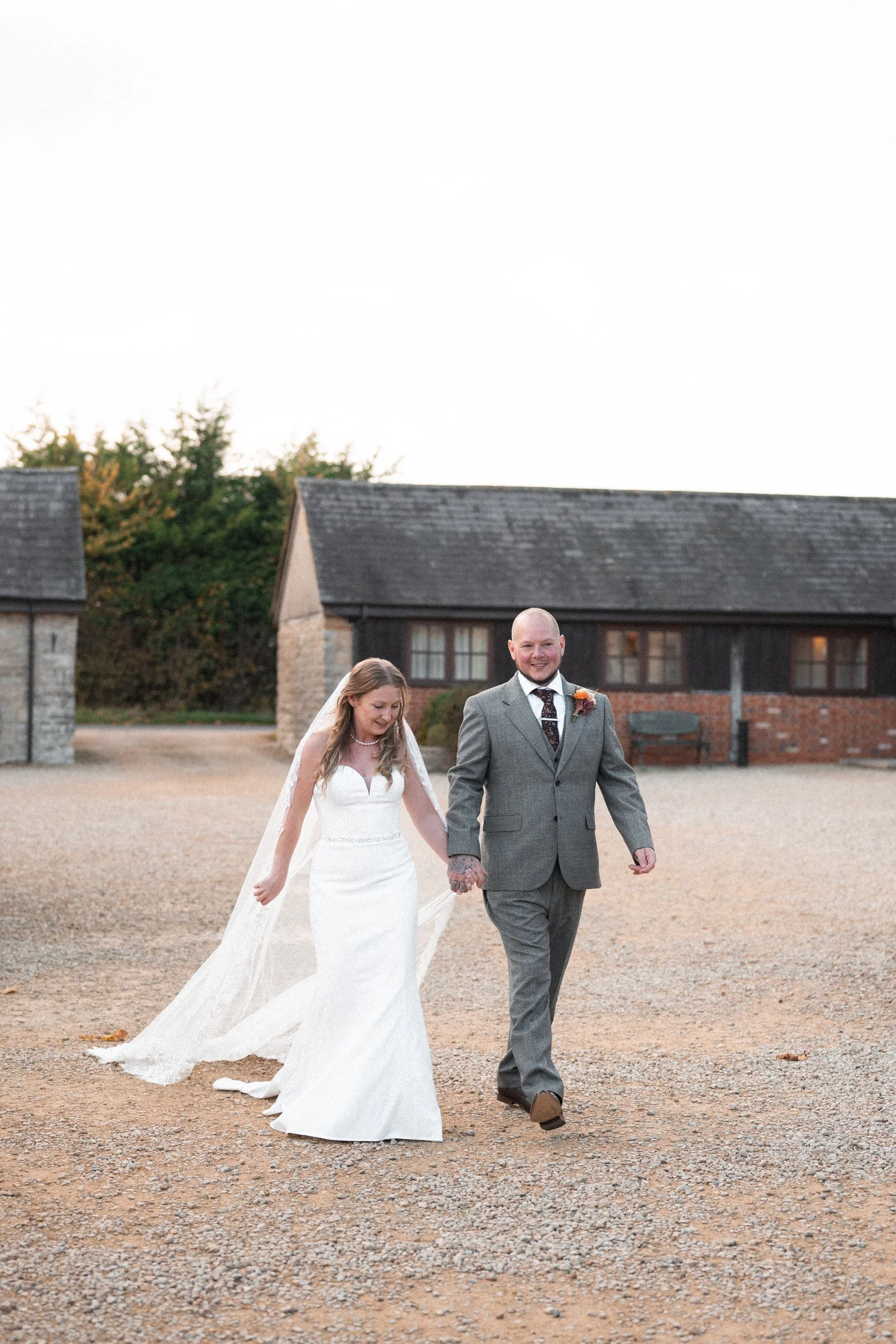 A bride and groom walking hand in hand outdoors during their wedding, smiling, with rustic barn buildings in the background.