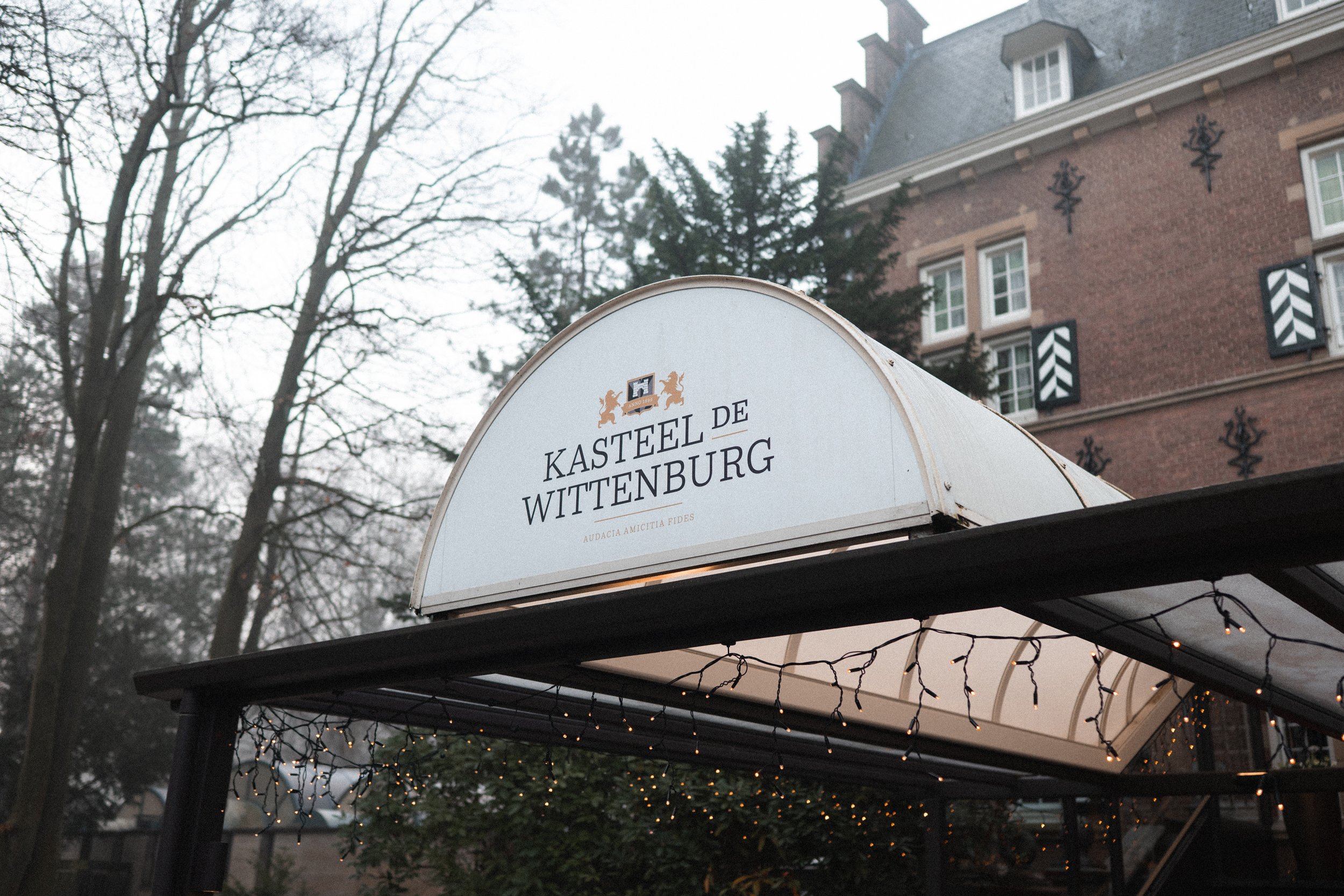Sign reading 'Kasteel de Wittenburg' with coat of arms above, attached to a black metal structure illuminated by small string lights, in front of a brick building with white window shutters and decorative black metal accents, trees with bare branches