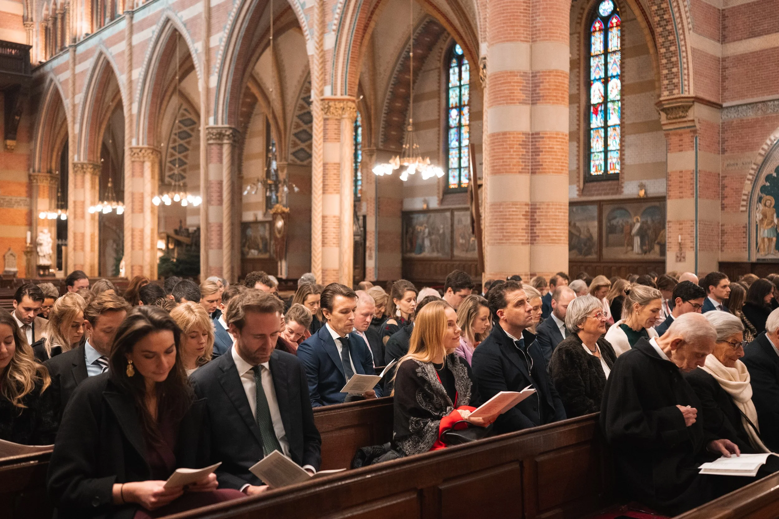 People sitting in pews inside a church, reading books or prayer materials, with stained glass windows and brick arches overhead.