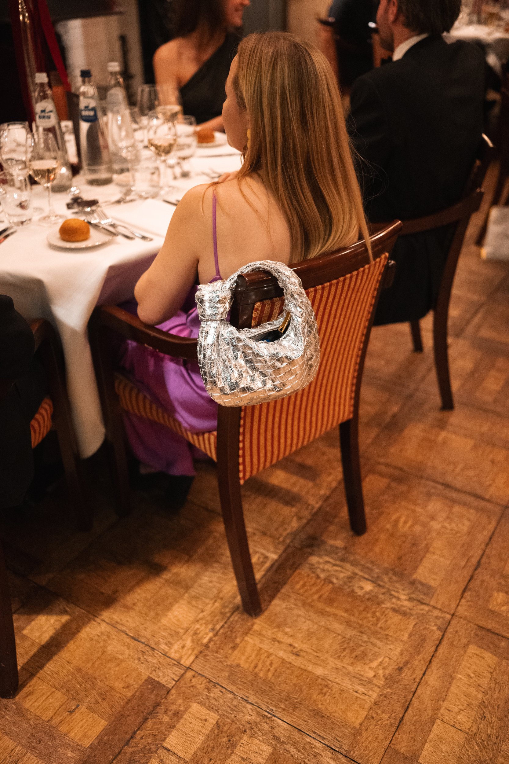 A woman sitting at a formal dinner table with a shiny silver handbag hanging on her chair.