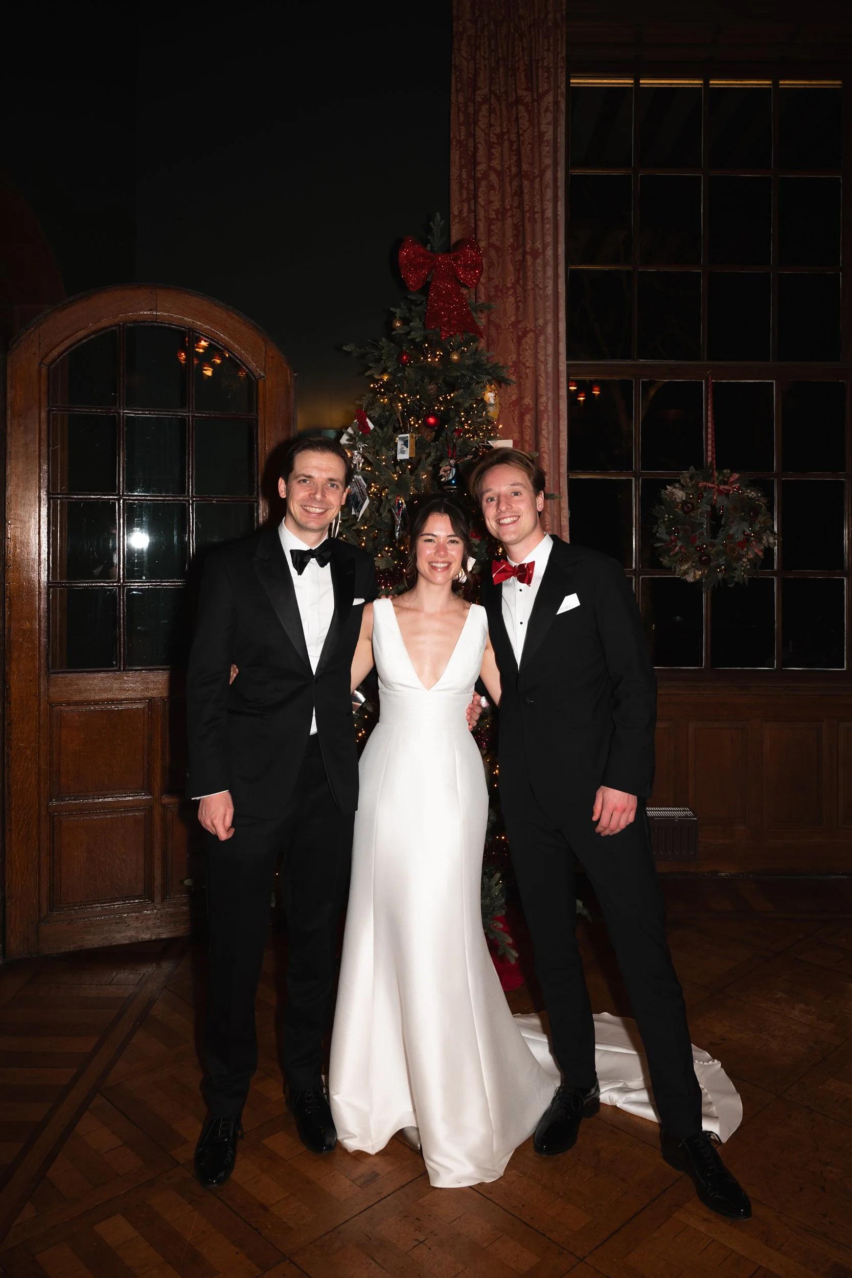 Three people dressed in formal wear (two men in tuxedos and a woman in a white gown) standing in front of a decorated Christmas tree, inside a wood-paneled room with large windows and floral curtains.