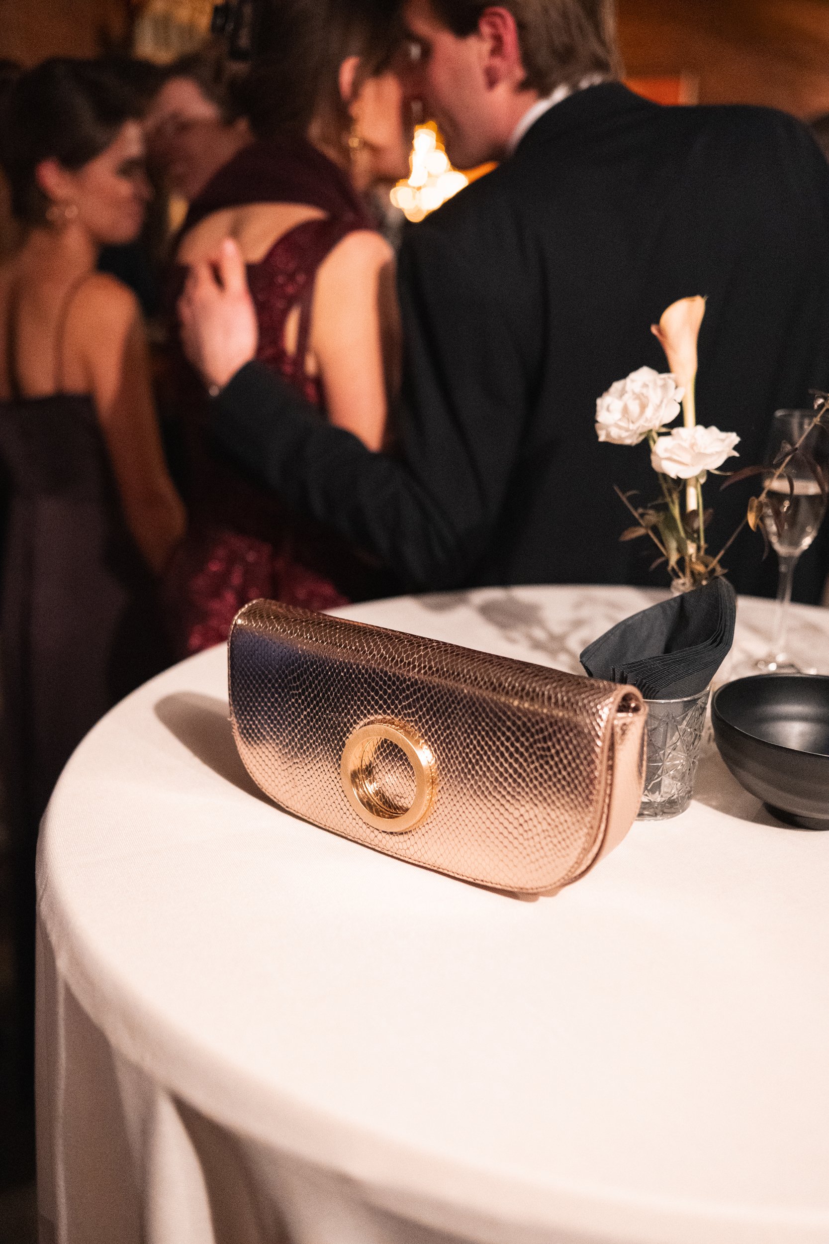 A round table with a rose gold clutch purse, a black bowl, a bouquet of white flowers, and a wine glass. In the background, people are mingling and dancing at a formal event.