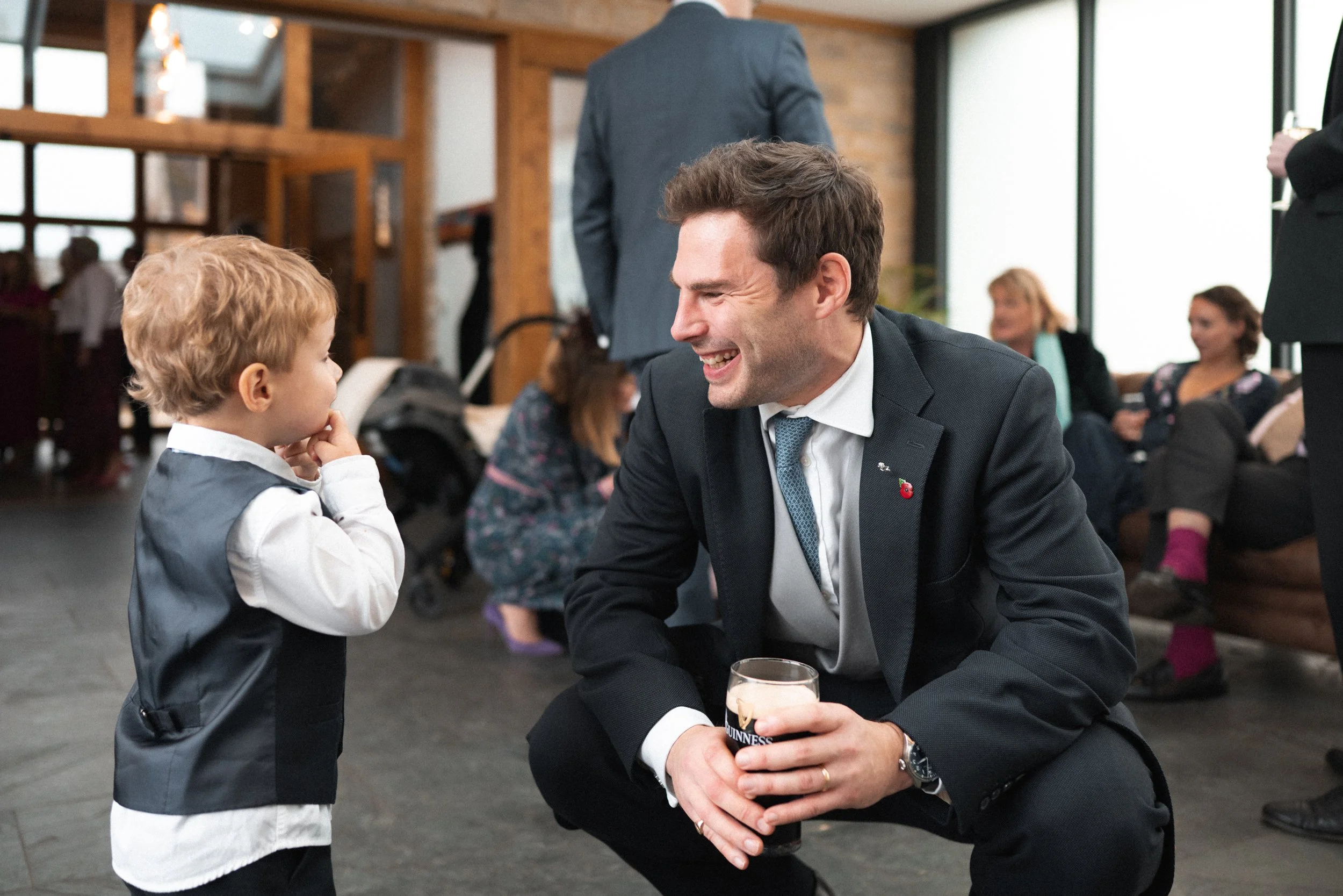 A man in a suit with a Guinness pint squats to talk to a young boy, who is wearing a vest and white shirt, in what appears to be a social gathering or party with other adults in the background.