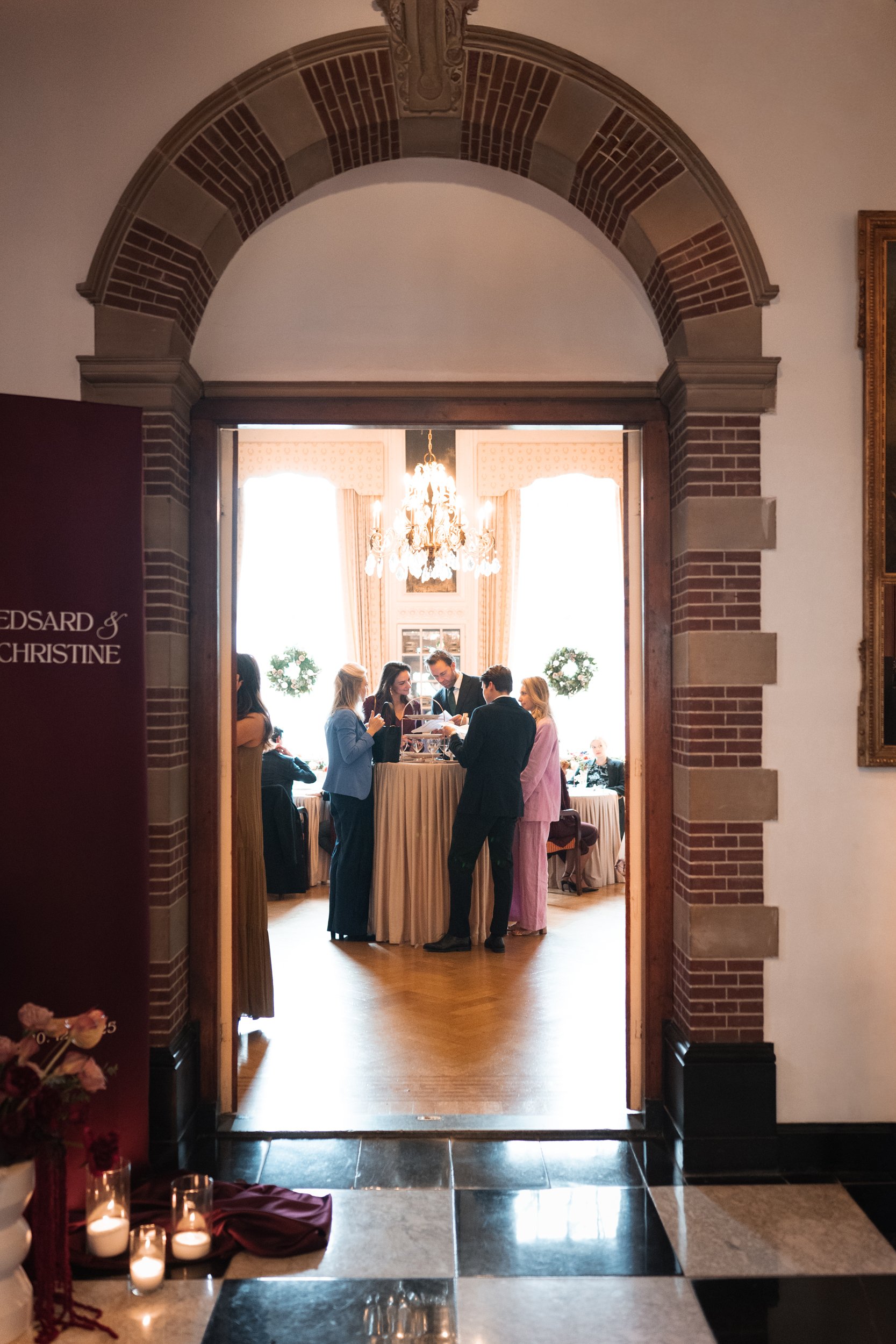 People gathered around a high-top table in a well-lit, elegant room with large windows and a chandelier, viewed through an arched doorway.