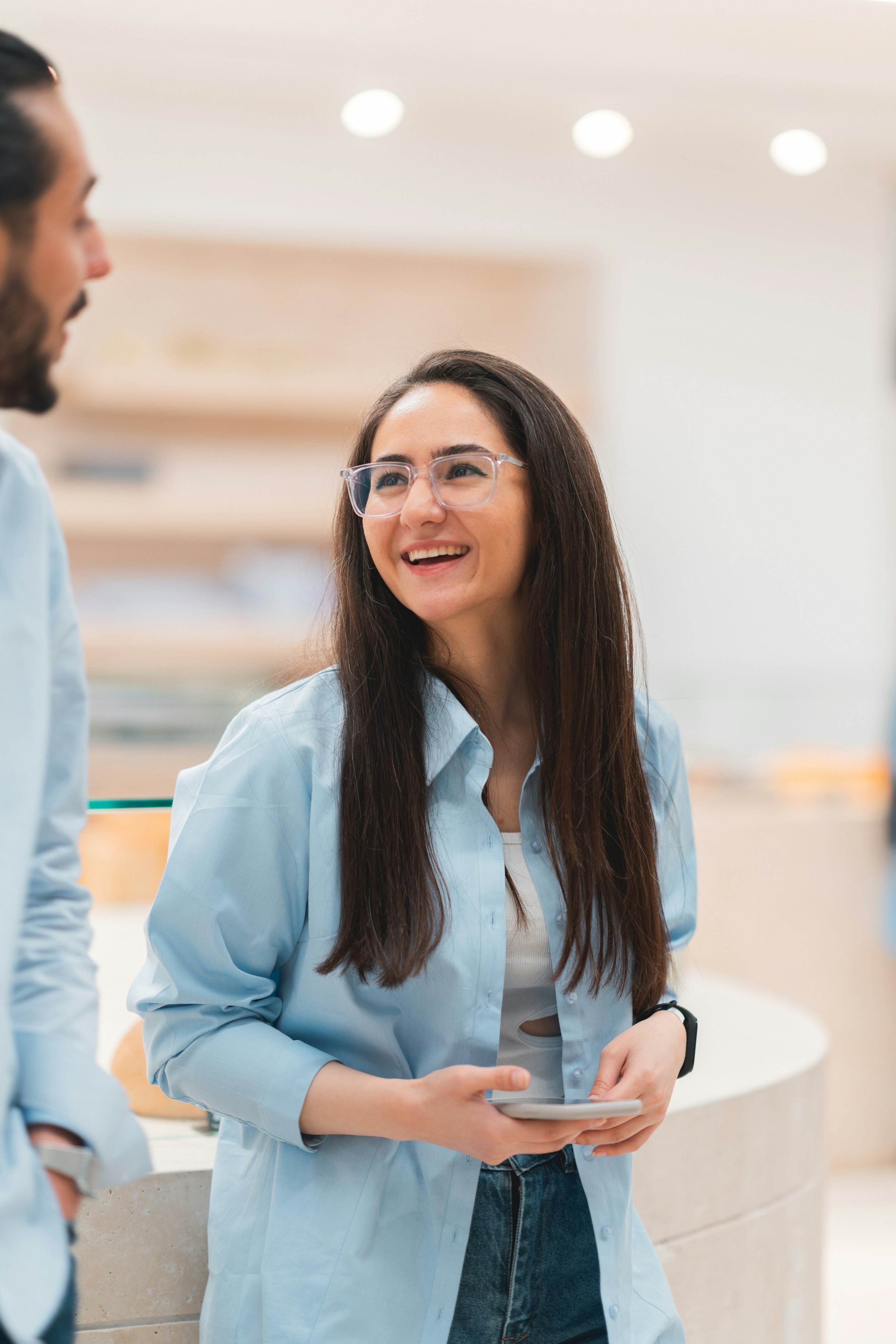Femme souriante portant des lunettes, tenant une tablette, discutant avec deux hommes dans un environnement de bureau.