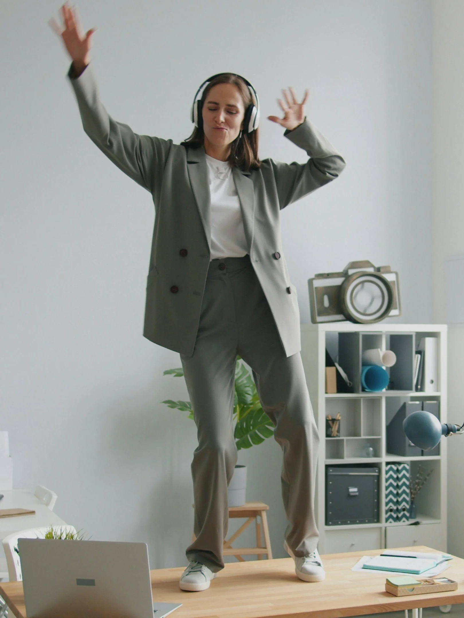 Une femme en costume décontracté, portant un casque audio, se tient debout sur une table de bureau, dans un environnement de bureau moderne avec étagères contenant des livres et des objets décoratifs, et un ordinateur portable sur la table.