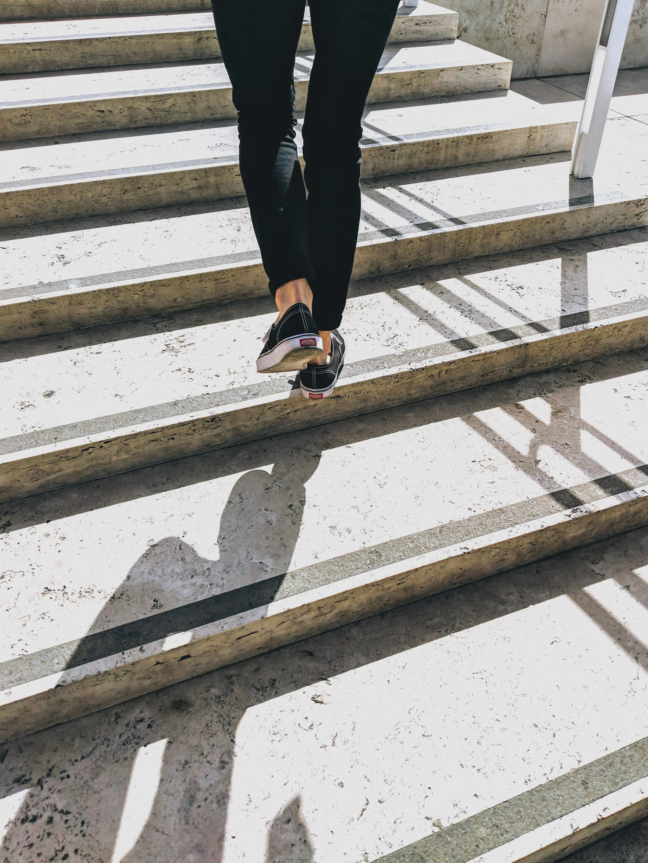 Une personne portant des chaussures Vans noires marche sur des marches en pierre, avec des ombres longues et marquées dues à la lumière solaire.