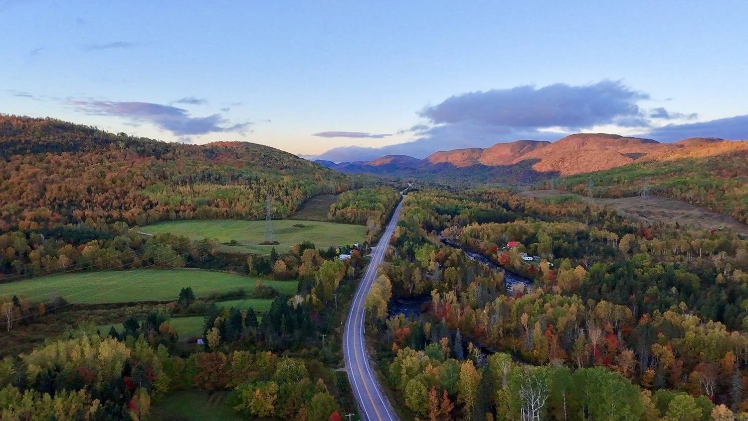 Paysage de montagnes avec forêt colorée en automne, une rivière, une route, quelques maisons, et un ciel partiellement nuageux.