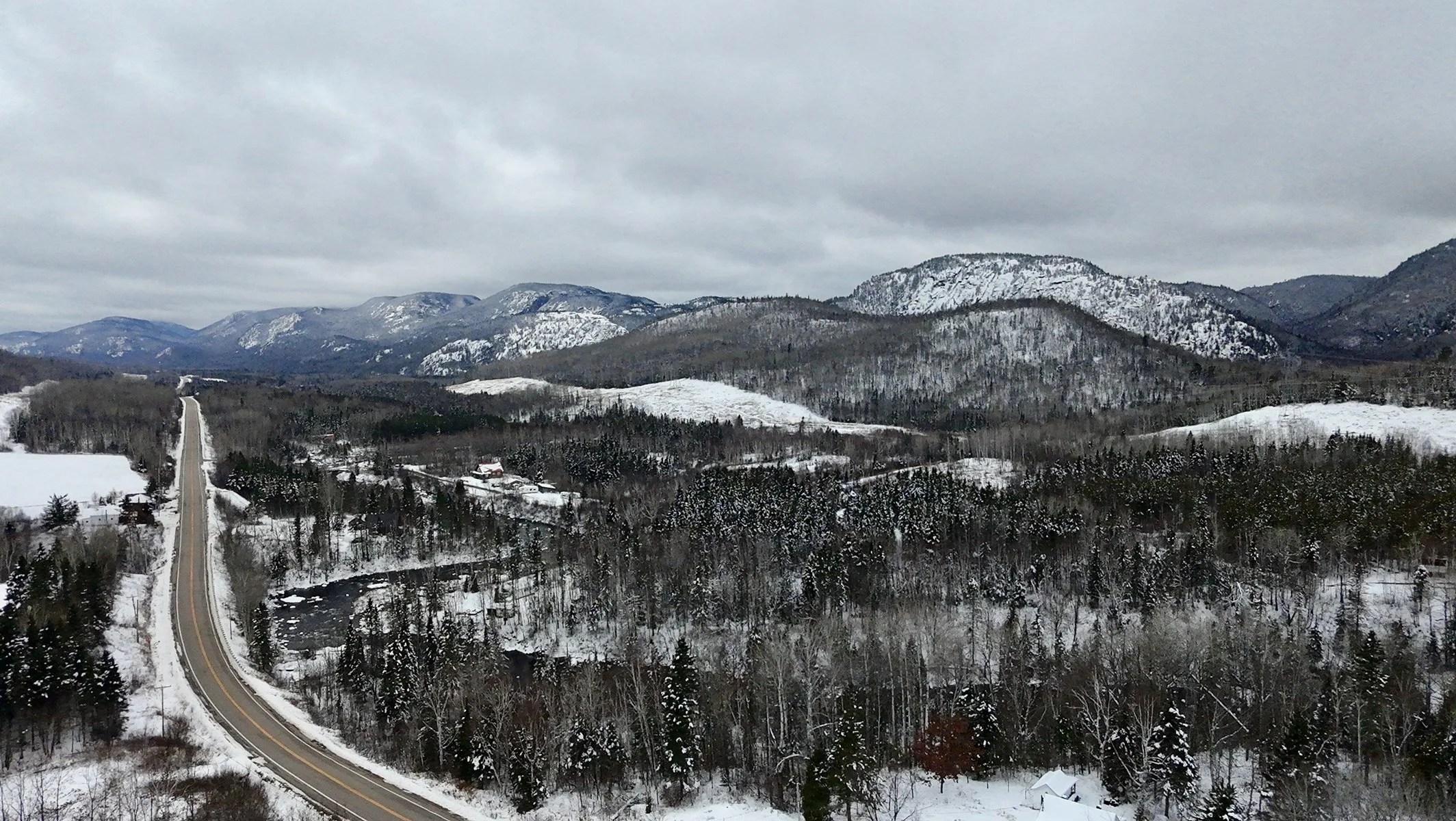 Terrains à vendre Petit-Saguenay. Paysage de montagnes enneigées, forêts et un chemin sinueux dans un environnement hivernal sous un ciel nuageux.