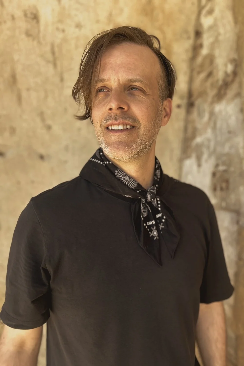David Pineault, fondateur d'Octave Immobilier, portant une chemise noire et un foulard noir avec un motif blanc, regardant vers la droite, avec un mur en pierre en arrière-plan.