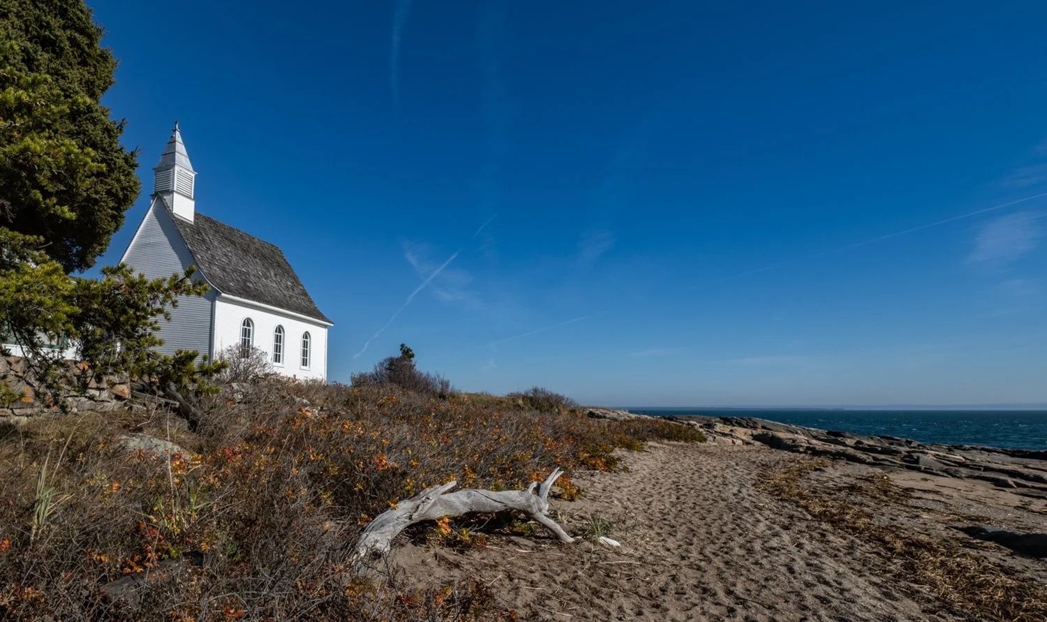 Une petite église blanche sous un ciel bleu, située près d'une plage avec du sable, des rochers et des buissons, à proximité de l'océan.