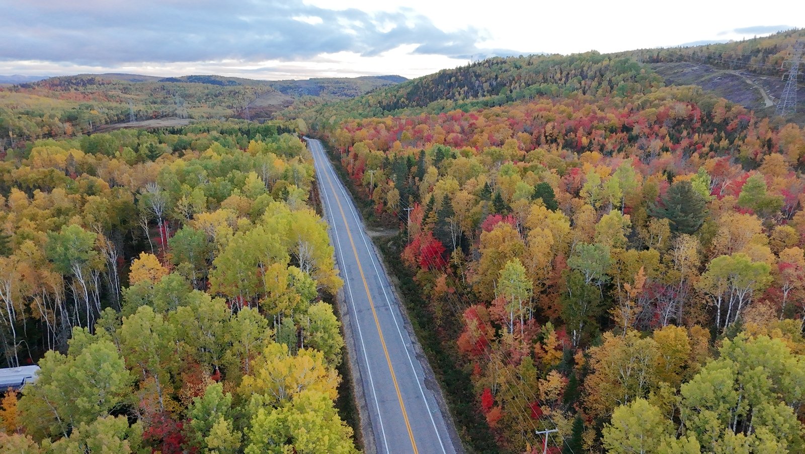 Vue aérienne d'une route traversant une forêt en automne avec des arbres colorés en rouge, jaune et vert, sous un ciel nuageux.