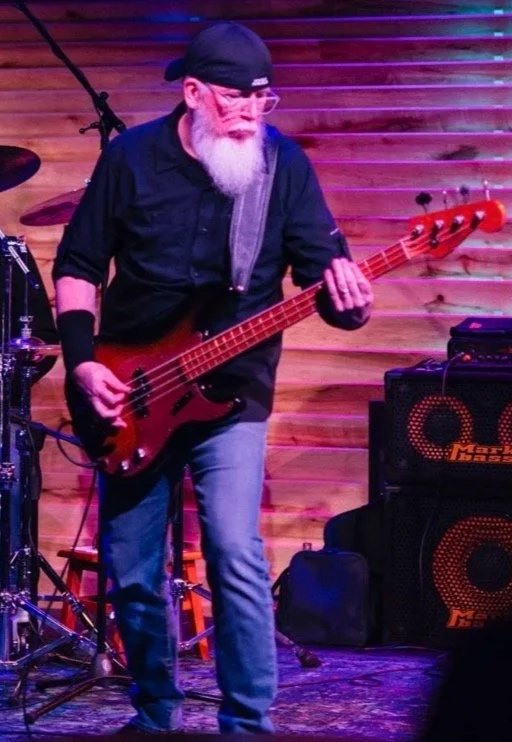 A man wearing sunglasses and a baseball cap playing electric guitar on stage with colorful lighting.