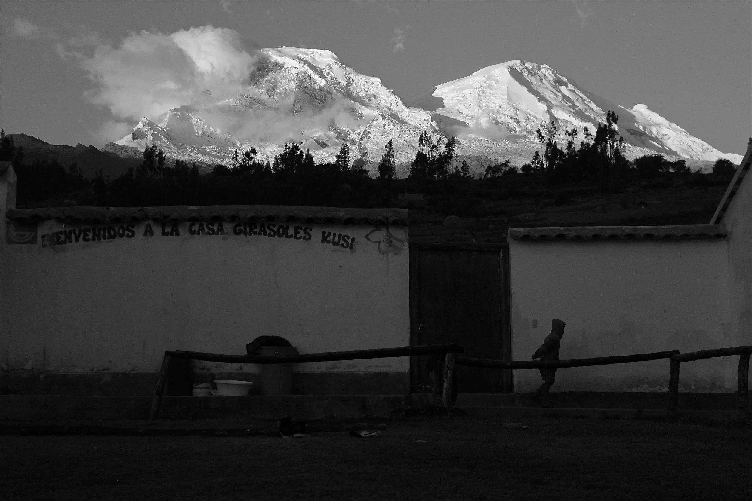 Black and white photo of a snow-capped mountain in the background with a small person walking near a wall in the foreground, and a sign that reads 'Bienvenidos a la casa girasoles Kusi'.