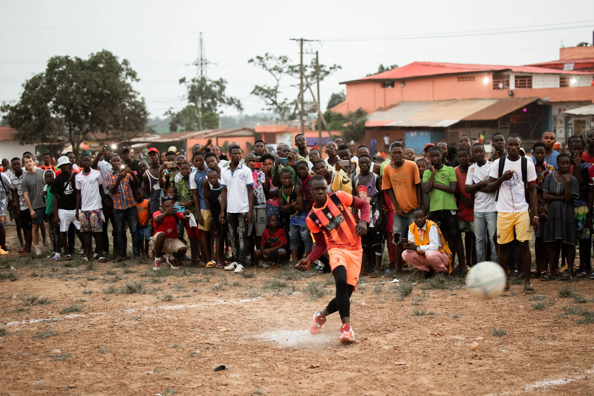 A young man in a striped orange and black soccer uniform kicks a soccer ball on a dirt field while a crowd of spectators watches behind him, with some recording the moment on their phones.