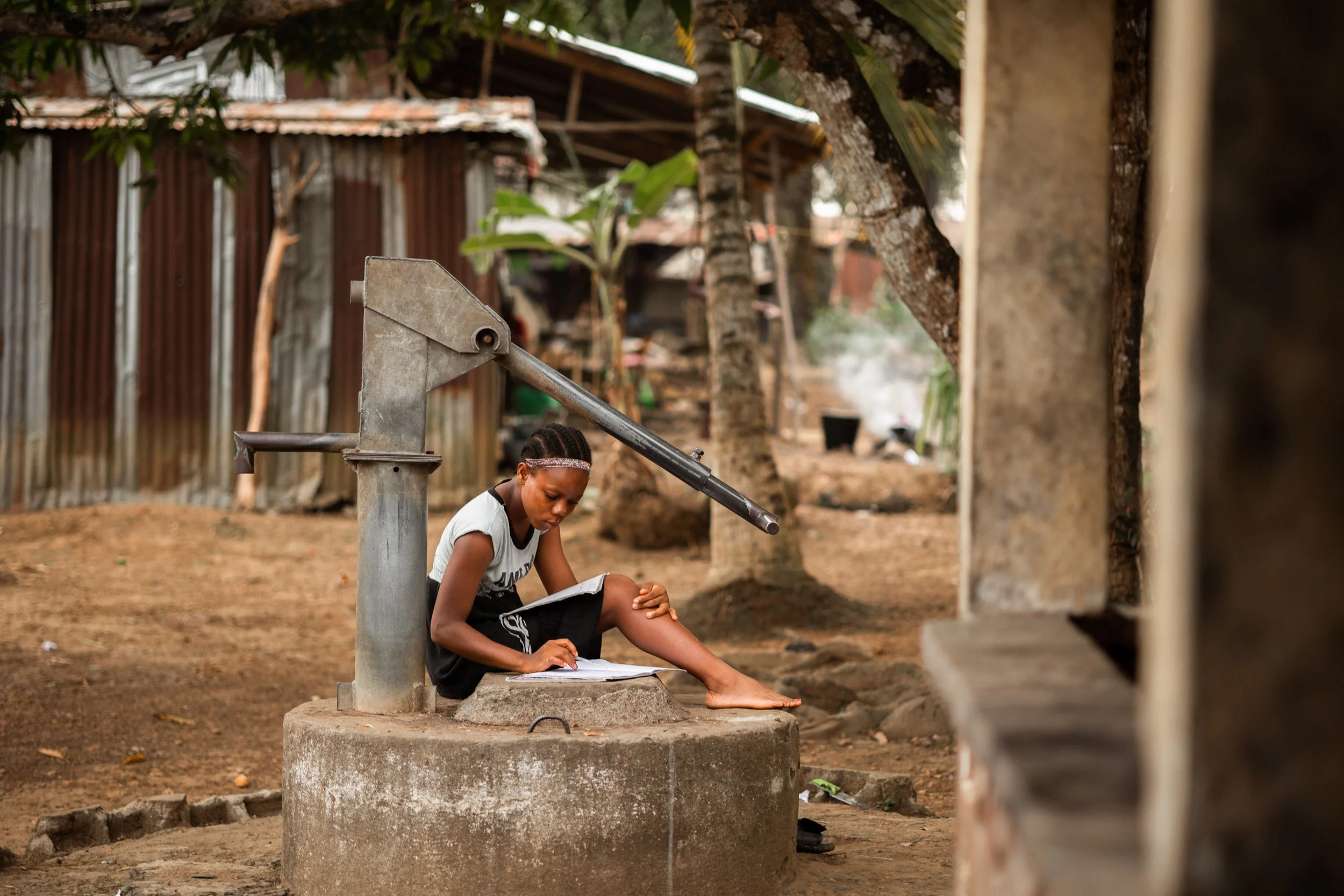 Young girl sitting on a concrete water pump in a rural area, writing or drawing on a piece of paper.
