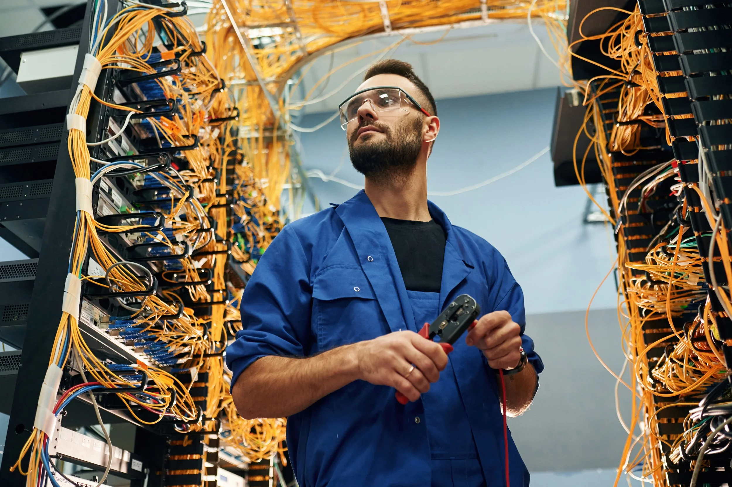 An electrician in safety glasses and blue coveralls working inside a data center, surrounded by server racks with numerous yellow and black cables.