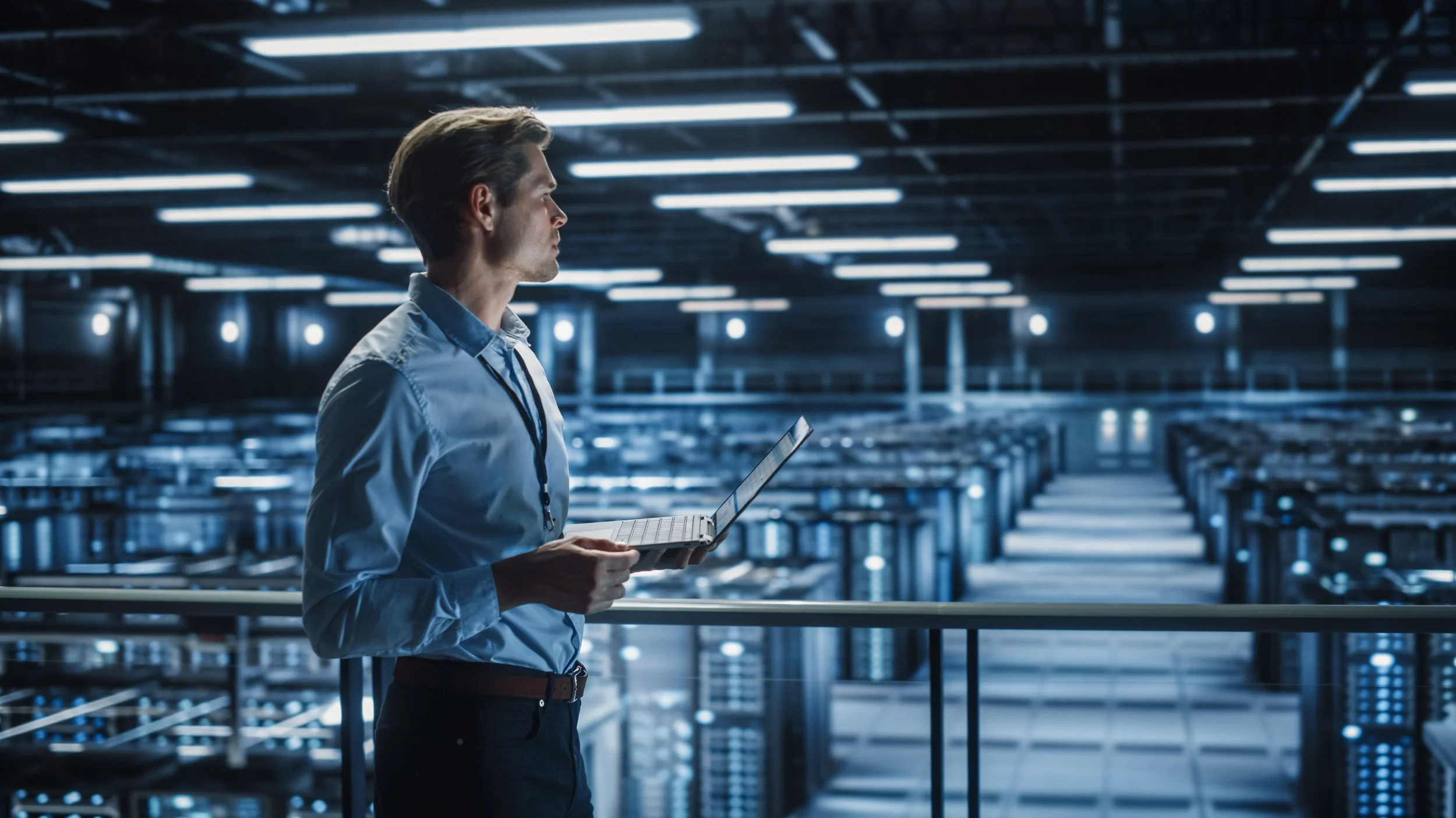 A man in a blue shirt holding a laptop, standing inside a large data center with rows of server racks and bright overhead lights.