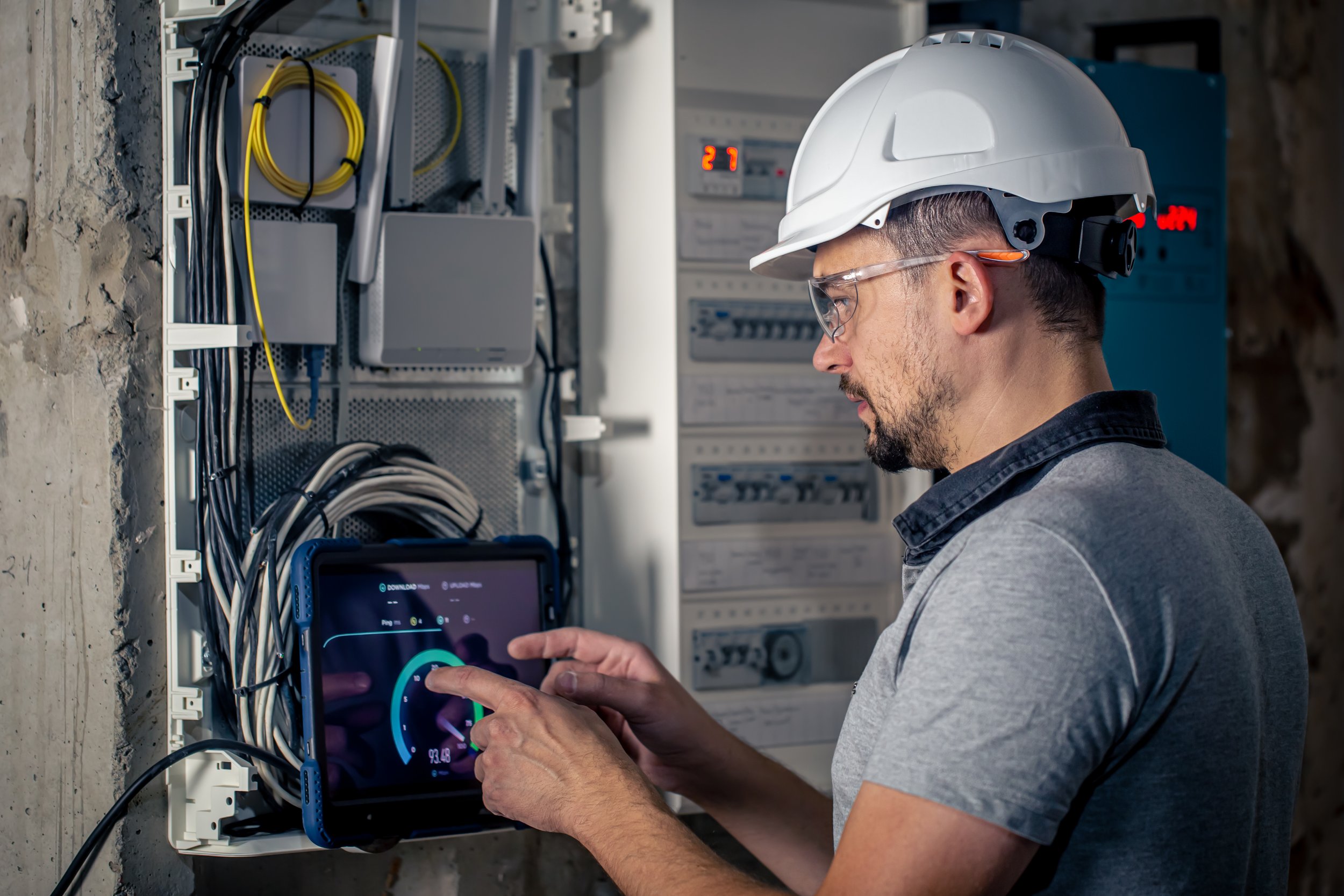 A technician wearing a white safety helmet and safety glasses using a tablet to inspect or troubleshoot an electrical control panel with numerous wires.