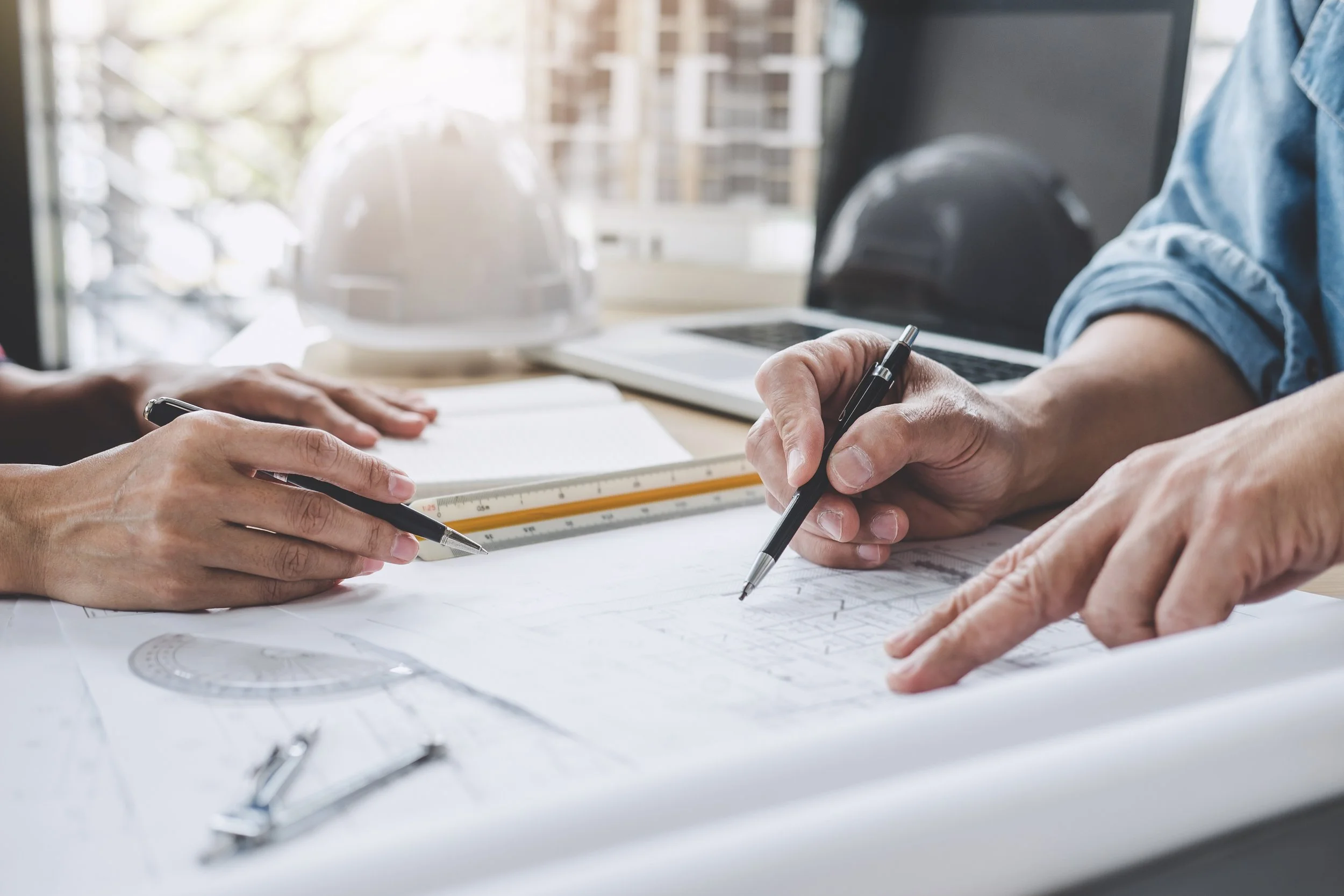 Two people discussing architectural plans, with hands pointing, holding pens, and a set square on the desk, with a safety helmet, laptop, and a window in the background.