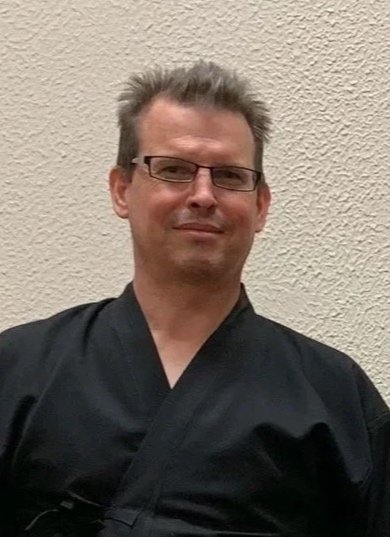 A man with glasses and gray hair, wearing a black martial arts uniform, standing against a beige textured wall.
