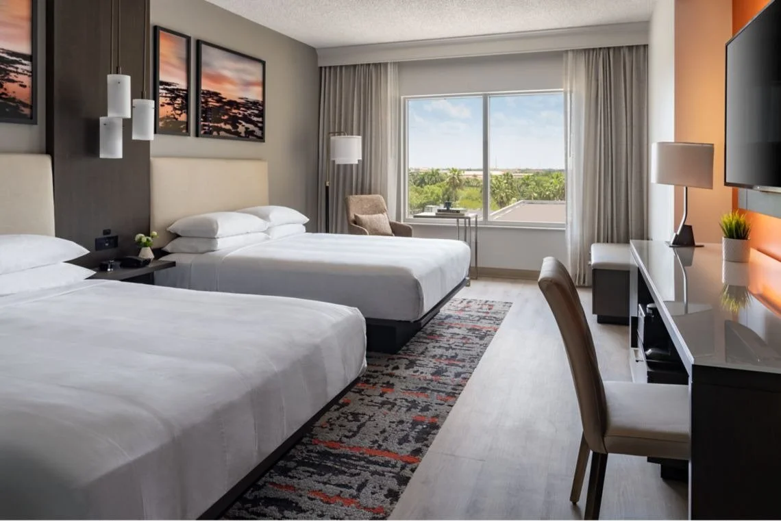 A modern hotel room with two white beds, a window overlooking greenery, a desk with a chair, and wall art at sunset.