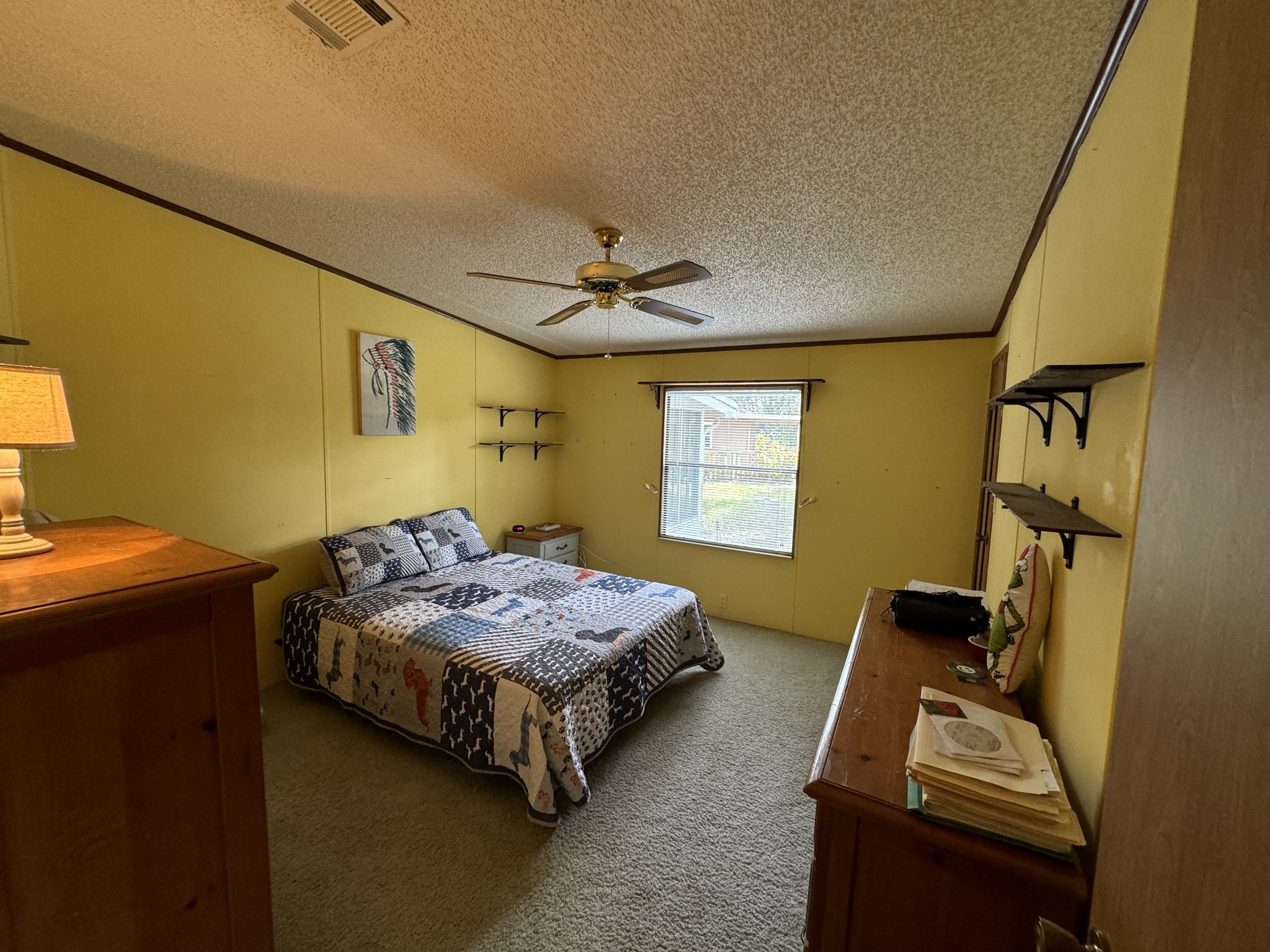 A bedroom with yellow walls and beige carpet. Contains a bed with a black and white patchwork quilt, a small white bedside table, a dresser with papers on top, a window with blinds, shelves on the wall, and a ceiling fan.
