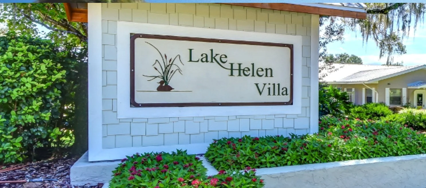Sign for Lake Helen Villa surrounded by green bushes and flowers, with a residential building in the background.
