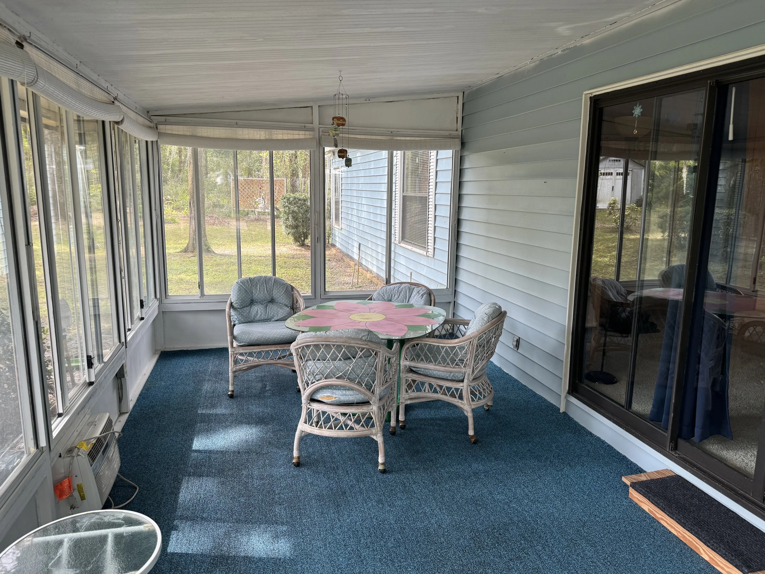 Enclosed porch with blue carpet, a round table with a floral design, five wicker chairs, large windows, and a view of the yard with trees and grass.