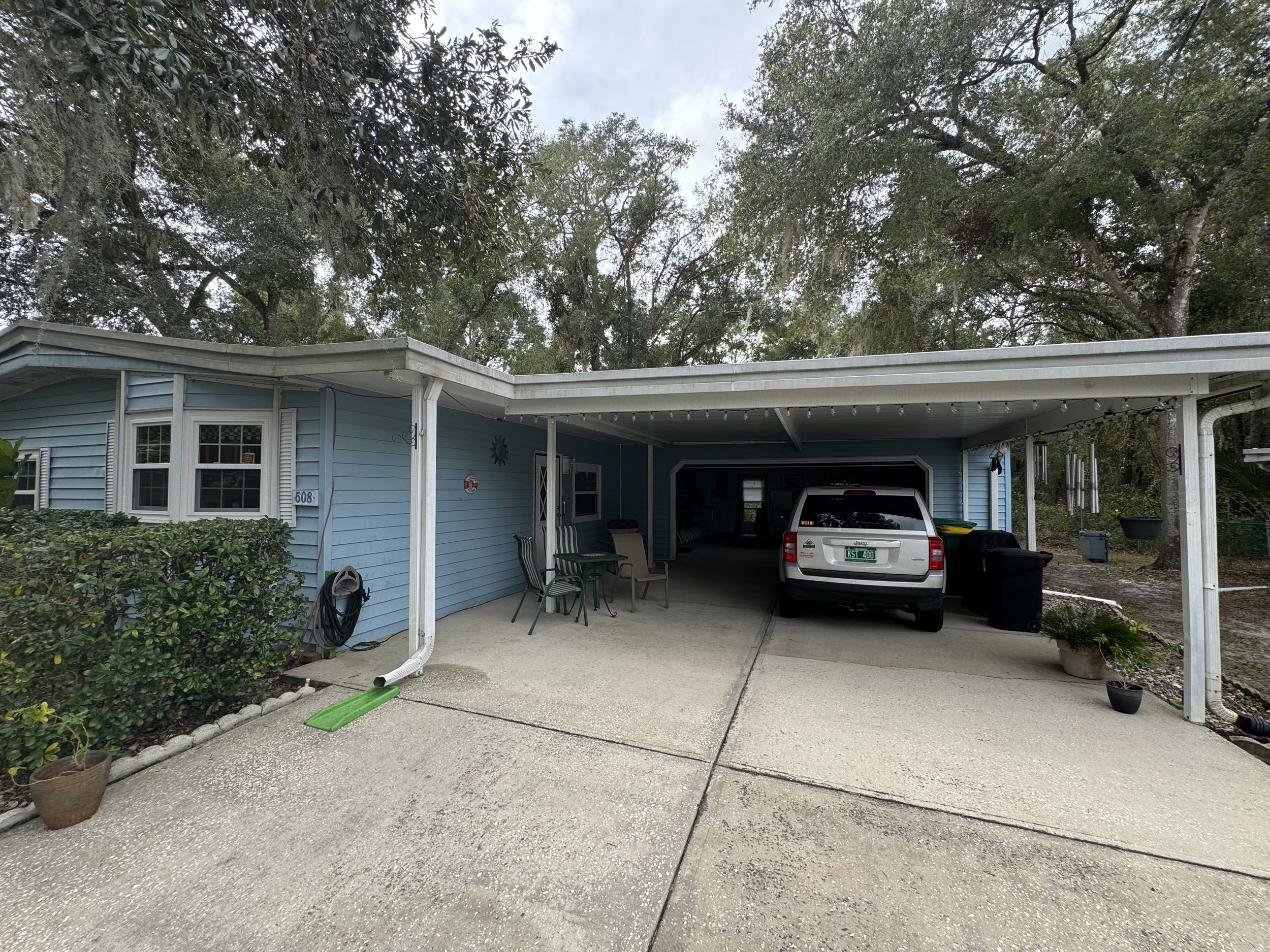 A house with a carport, blue siding, and a white SUV parked underneath. There are outdoor chairs, potted plants, and a garden hose on the side. Trees are visible in the background.