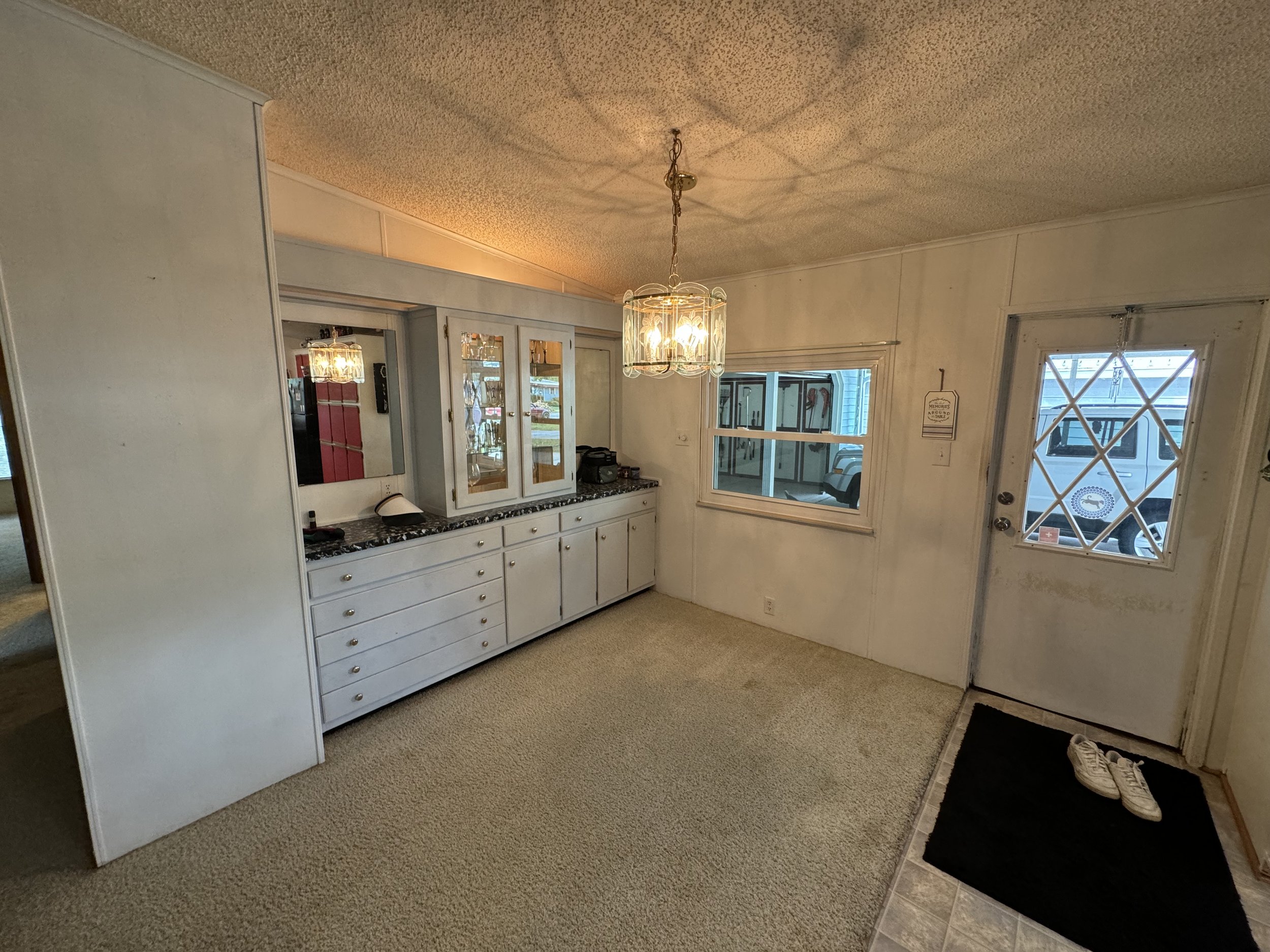Interior view of a dining area with a white built-in cabinet, a window, and a door with a diamond-patterned window, with sneakers on a black mat in the corner. A chandelier hangs from the ceiling.