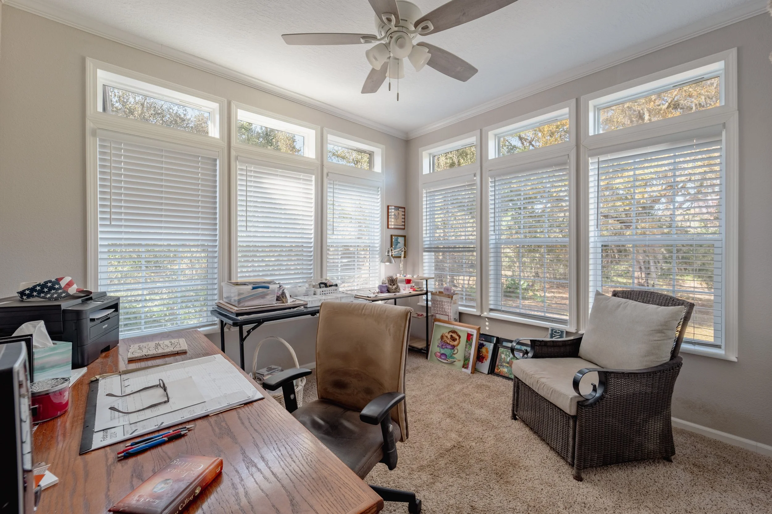 Home office with a large window, beige carpet, desk with papers and office supplies, a brown office chair, a wicker chair with a pillow, ceiling fan, and various artwork leaning against the wall.