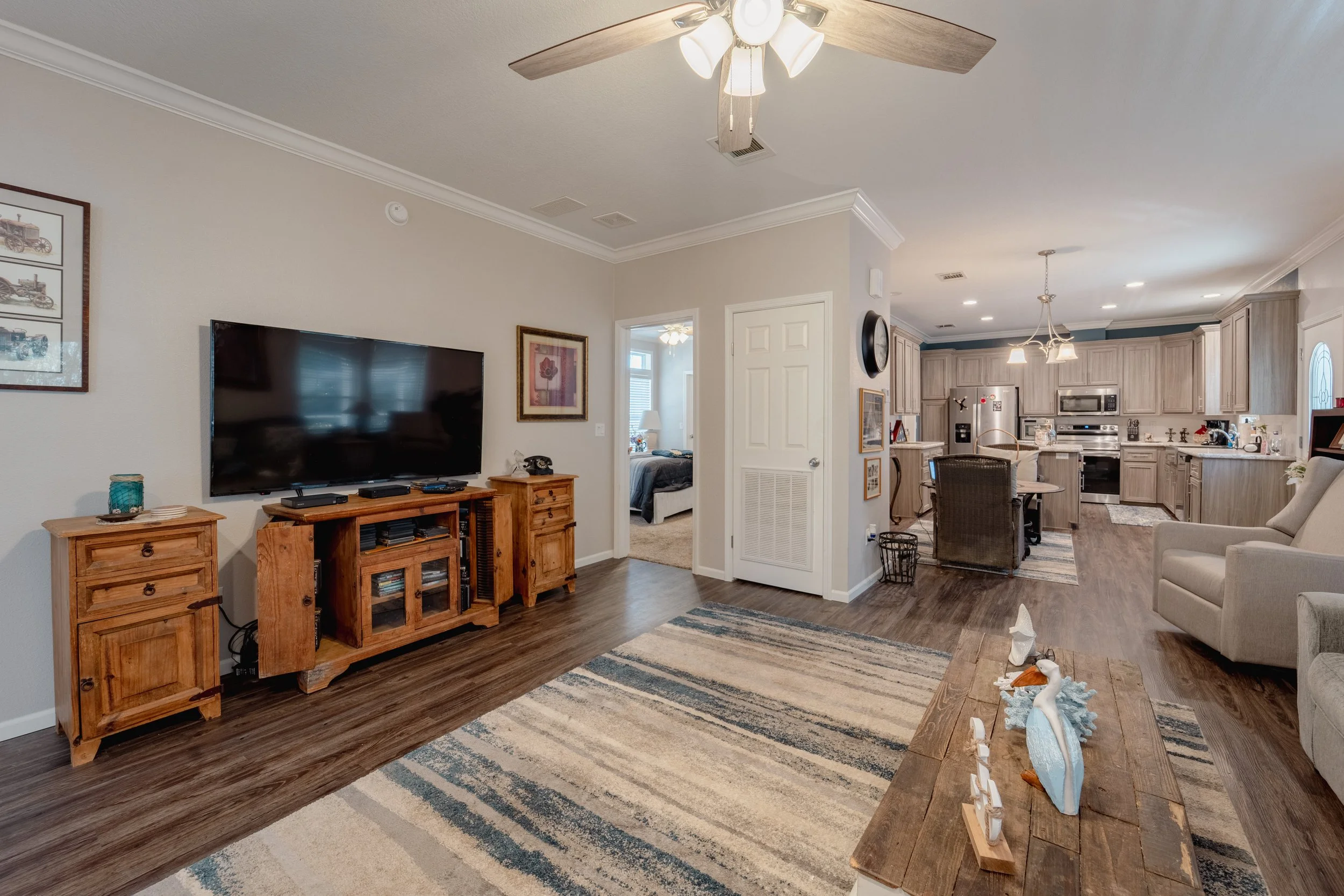 Living room with wooden TV stand, large flat-screen TV, beige walls, hardwood flooring, area rug, and view into kitchen and dining area.