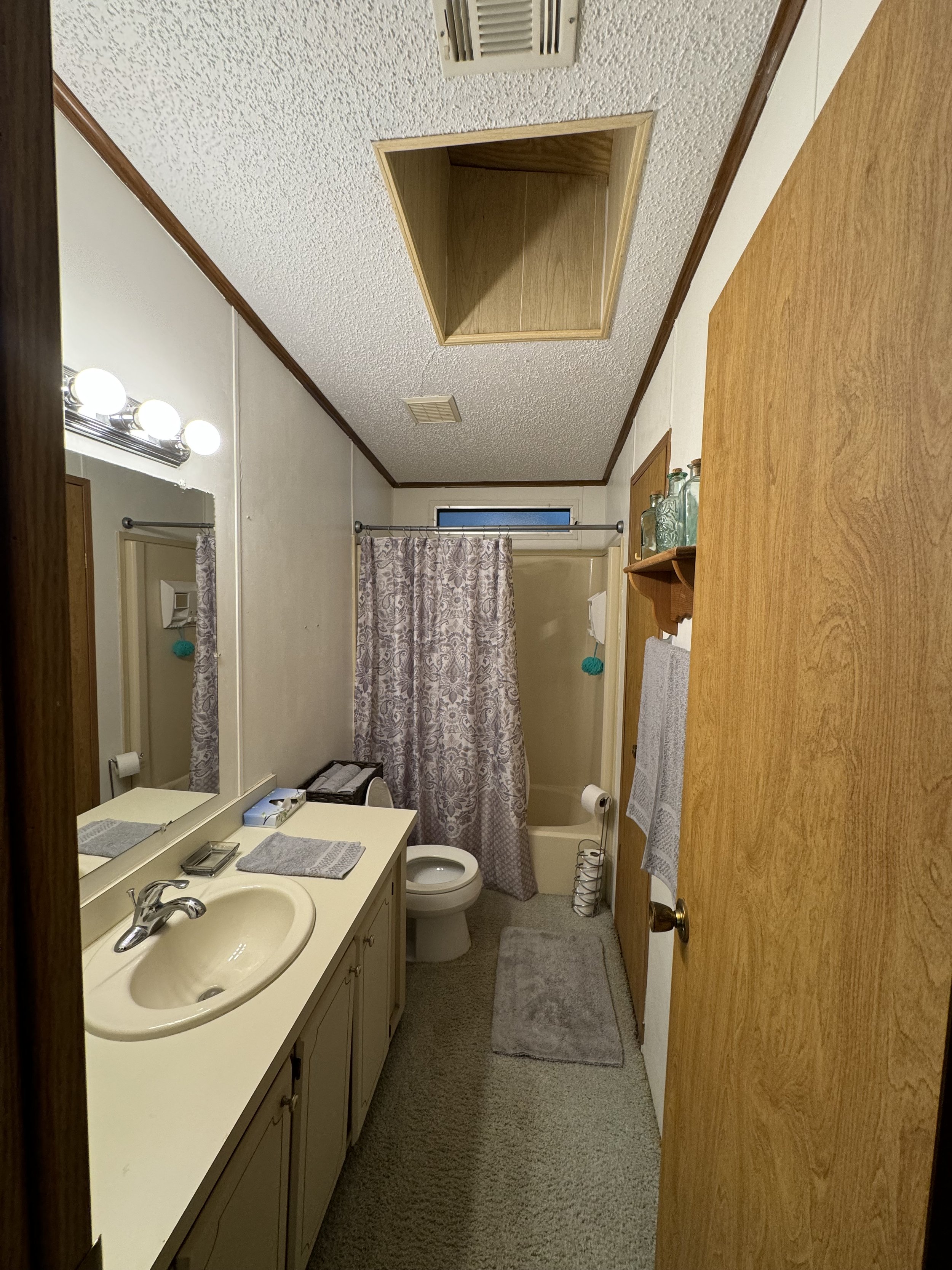 A small bathroom with a sink, mirror, toilet, bathtub with shower, wooden shelf with decorative glass bottles, ceiling vent, open ceiling hatch, rug, towel, and a window above the bathtub.