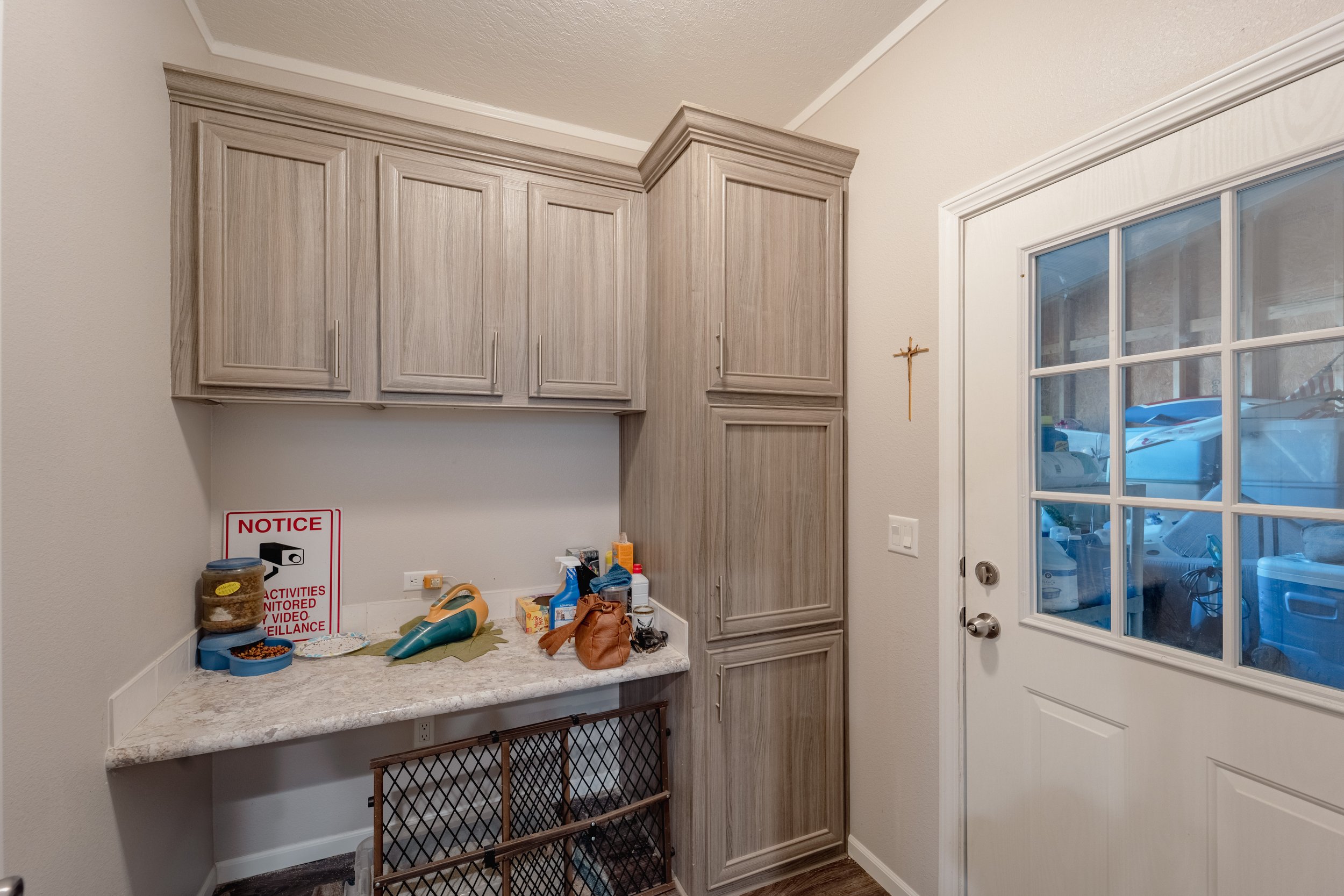 A laundry room with beige cabinets, a countertop with various items, a small wall cross, and a white door with a window showing a garage with cars.