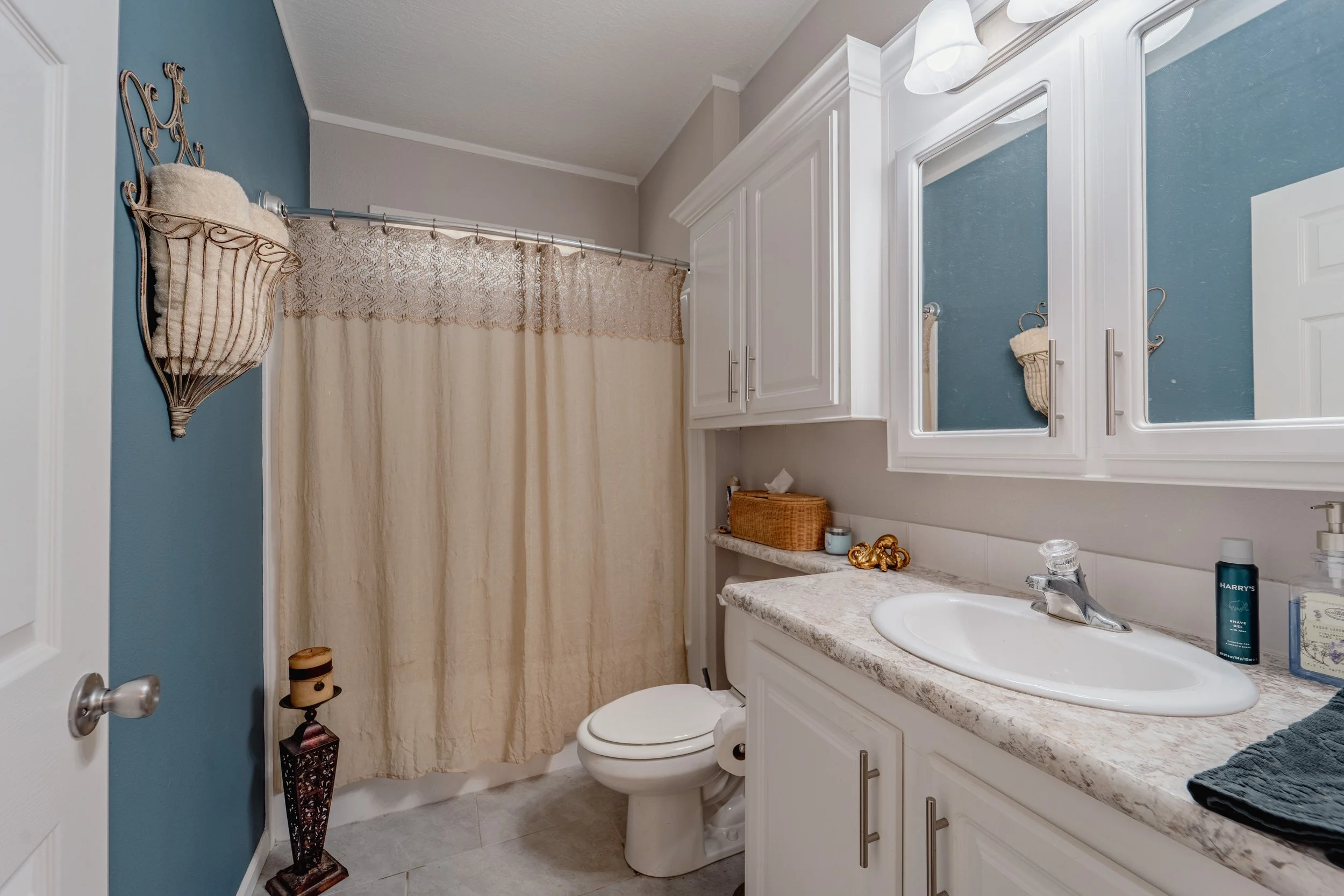 Bathroom with a blue accent wall, white vanity with sink, mirror cabinets, beige shower curtain, toilet, and decorative items on the countertop and wall