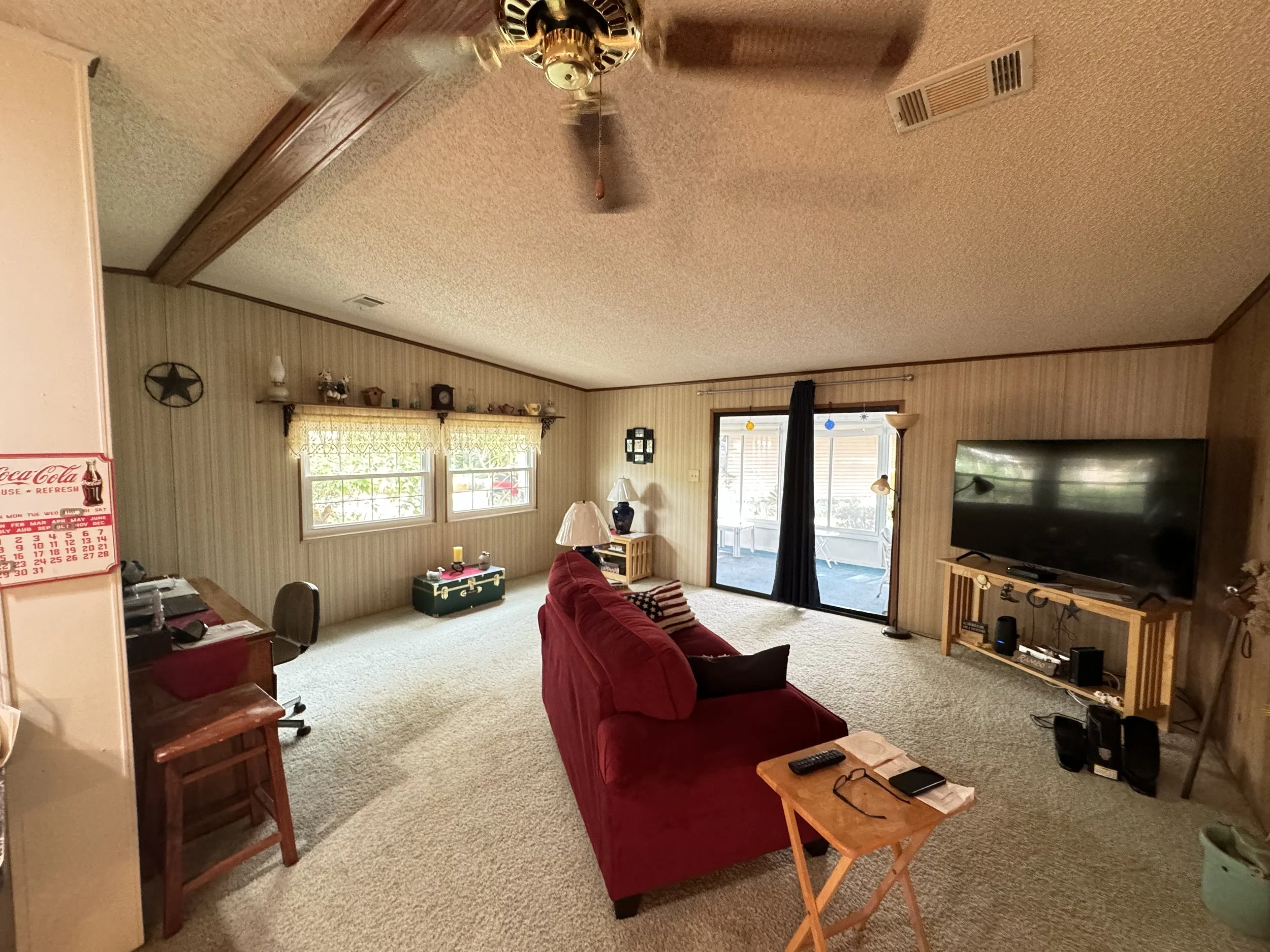 A living room with beige carpet, a red couch, a small wooden coffee table, and a large flat-screen TV on a wooden stand. There are two windows with yellow curtains, a sliding glass door with a black curtain, and various decorative items on shelves an