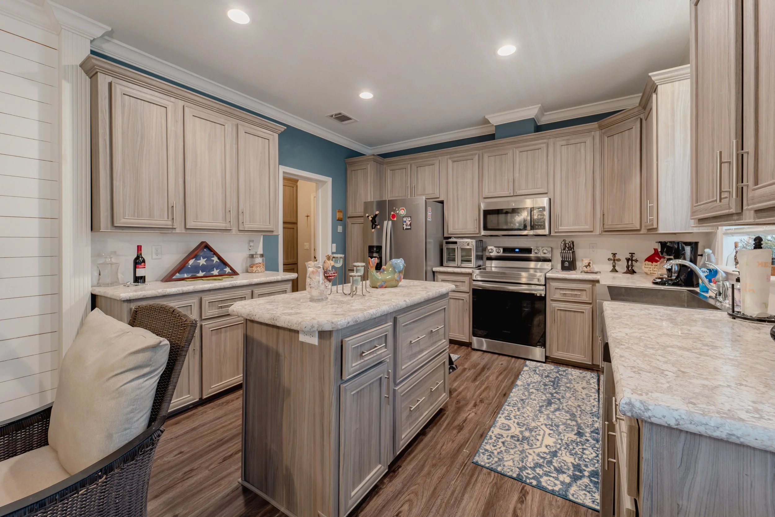 Kitchen with light wood cabinets, stainless steel appliances, granite countertops, and a blue accent wall. A kitchen island and a small patterned rug are also visible.