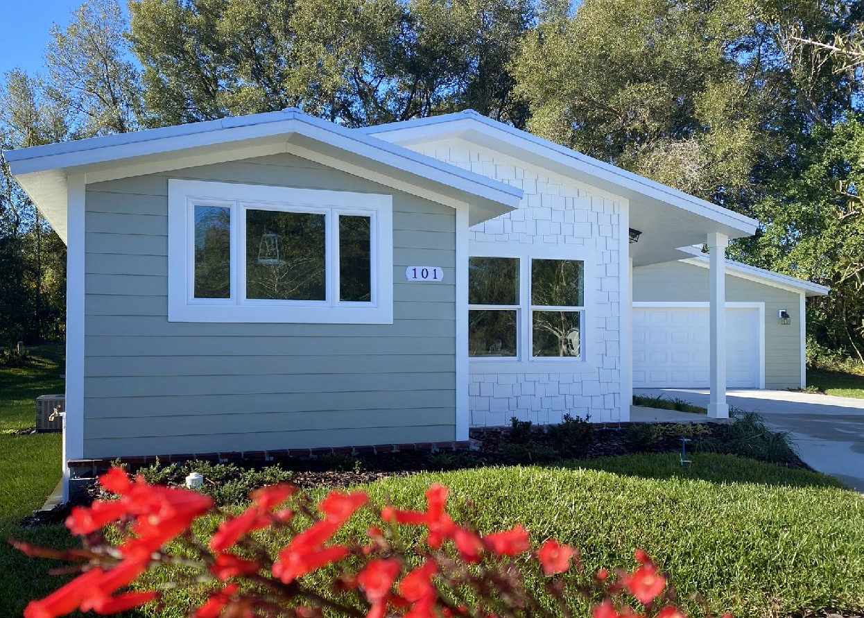 Front view of a modern, light-colored house with a garage, surrounded by grass, trees, and a small shrub with red flowers in the foreground.