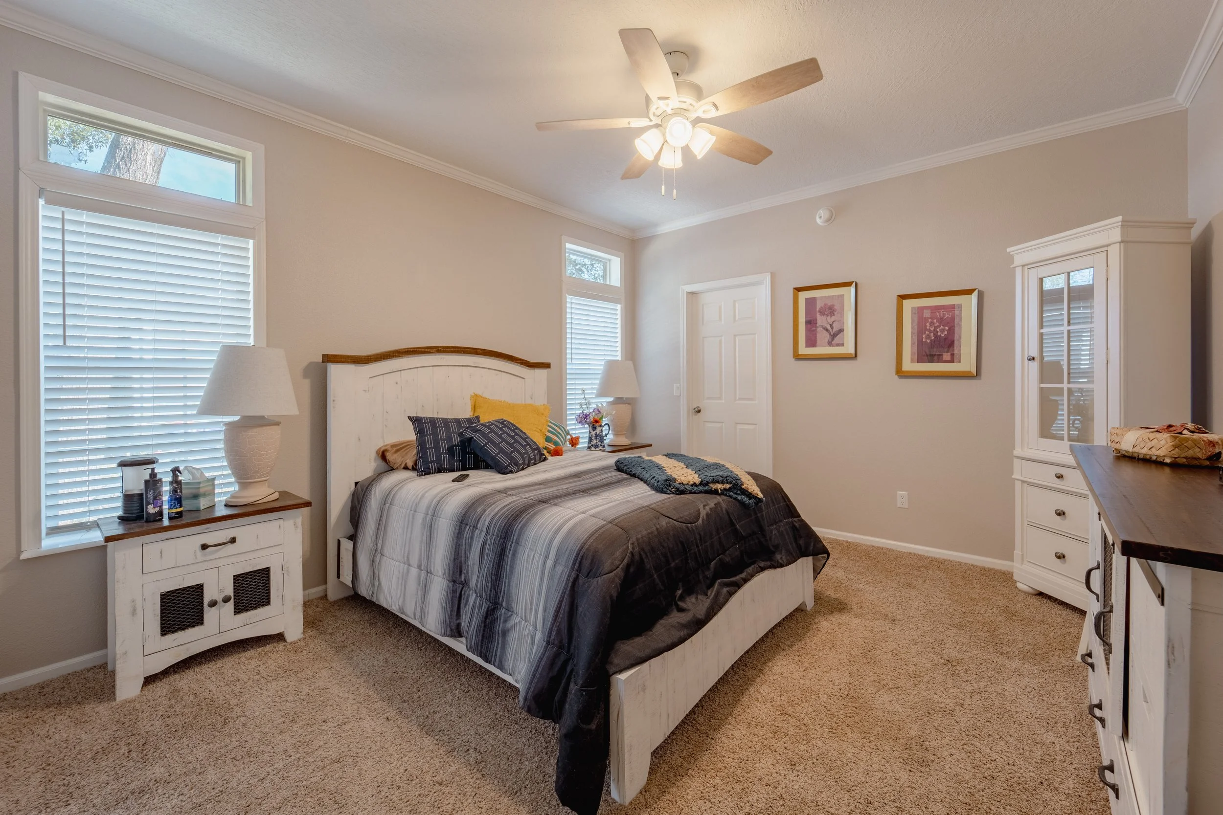 Bedroom with a white bed, dark bedding, and pillows, two nightstands with lamps and decorative items, beige walls with two framed pink artwork pieces, white dresser, and a white cabinet, beige carpet, ceiling fan, and natural light from multiple wind