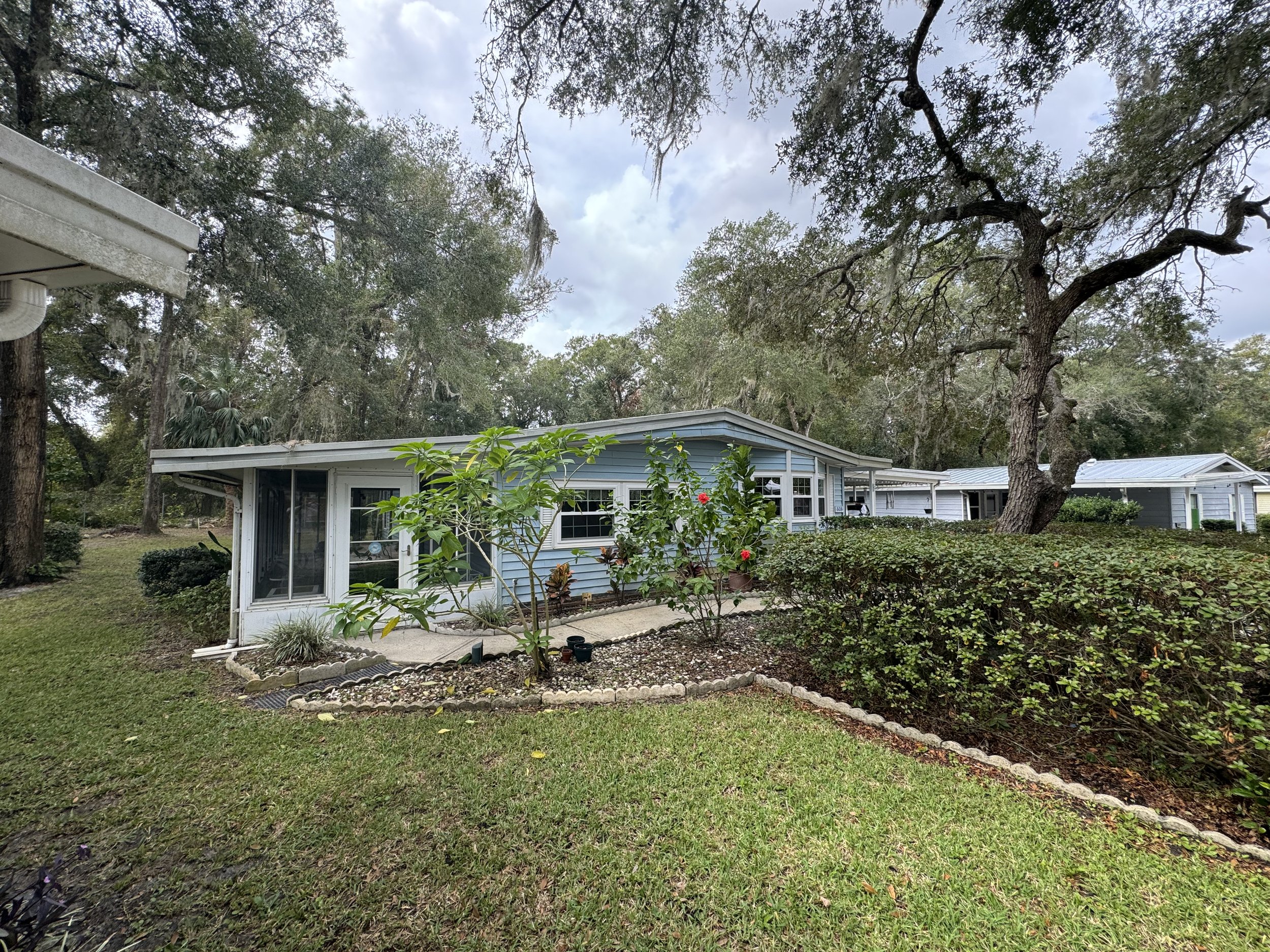 A suburban house with blue siding, surrounded by trees and a garden with bushes and flowering plants, under a cloudy sky.