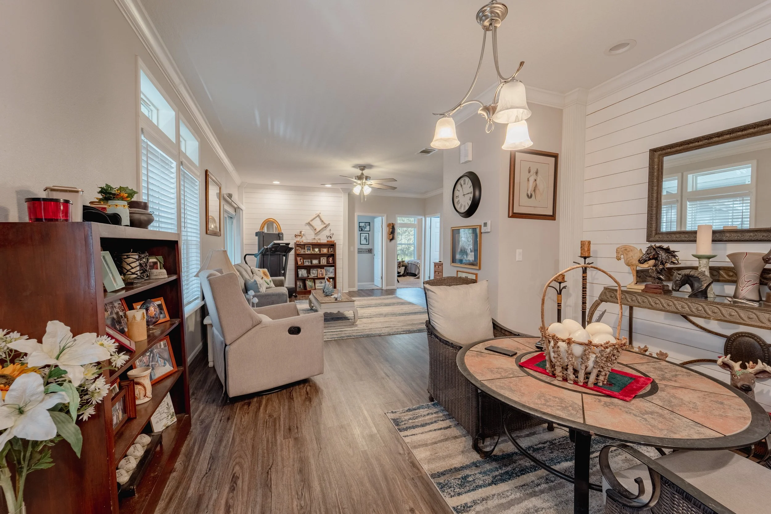 Living room and dining area with beige and white walls, wooden flooring, a large mirror, a wooden bookshelf, a sofa, armchairs, a treadmill in the background, various pictures and decorations on the walls, a ceiling fan, a chandelier, and a striped a