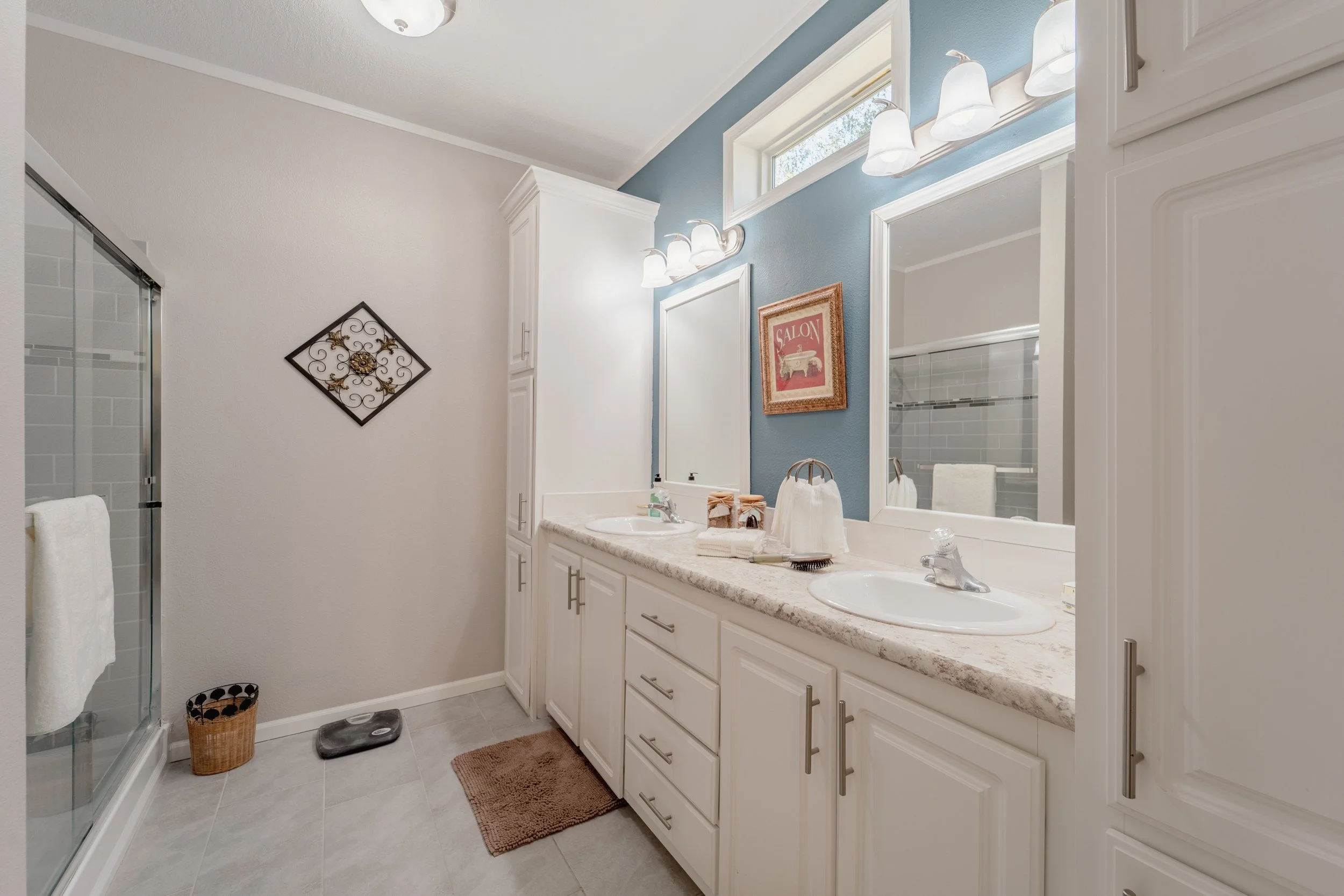 Bathroom with dual sinks, white cabinets, mirror, wall-mounted light fixtures, shower with glass door, towel, scale, and wall art.