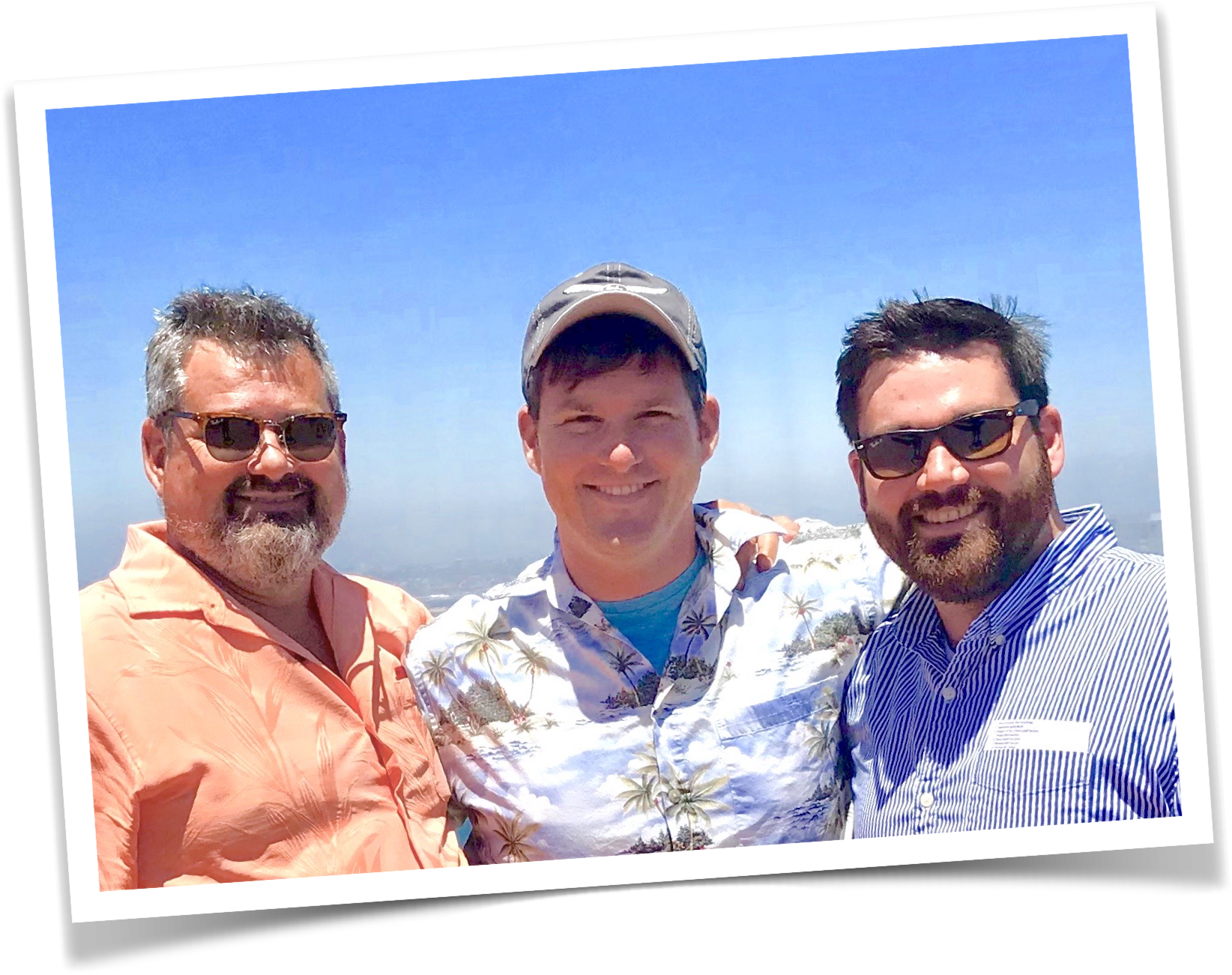 Three men smiling outdoors on a sunny day, wearing casual shirts and sunglasses, with a clear blue sky in the background.