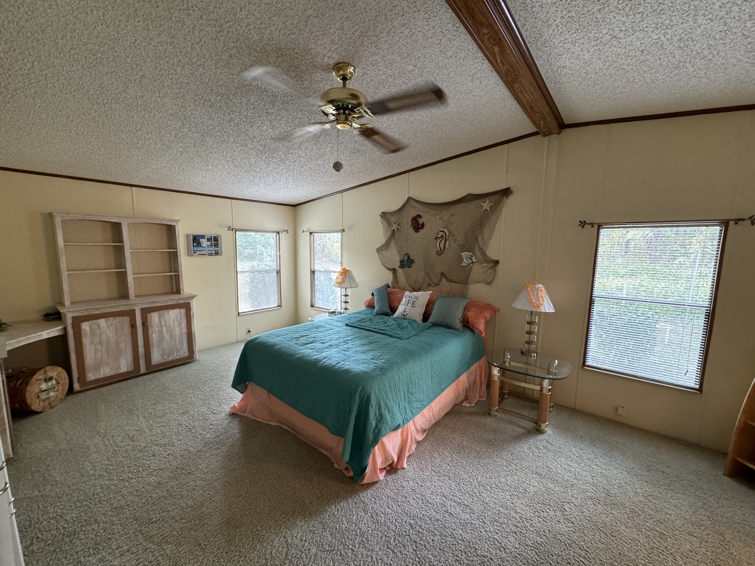 A bedroom with a bed, teal and pink bedding, and seaside-themed wall decor. Side tables with lamps on either side of the bed, windows with blinds, a ceiling fan, and an empty wooden bookcase.