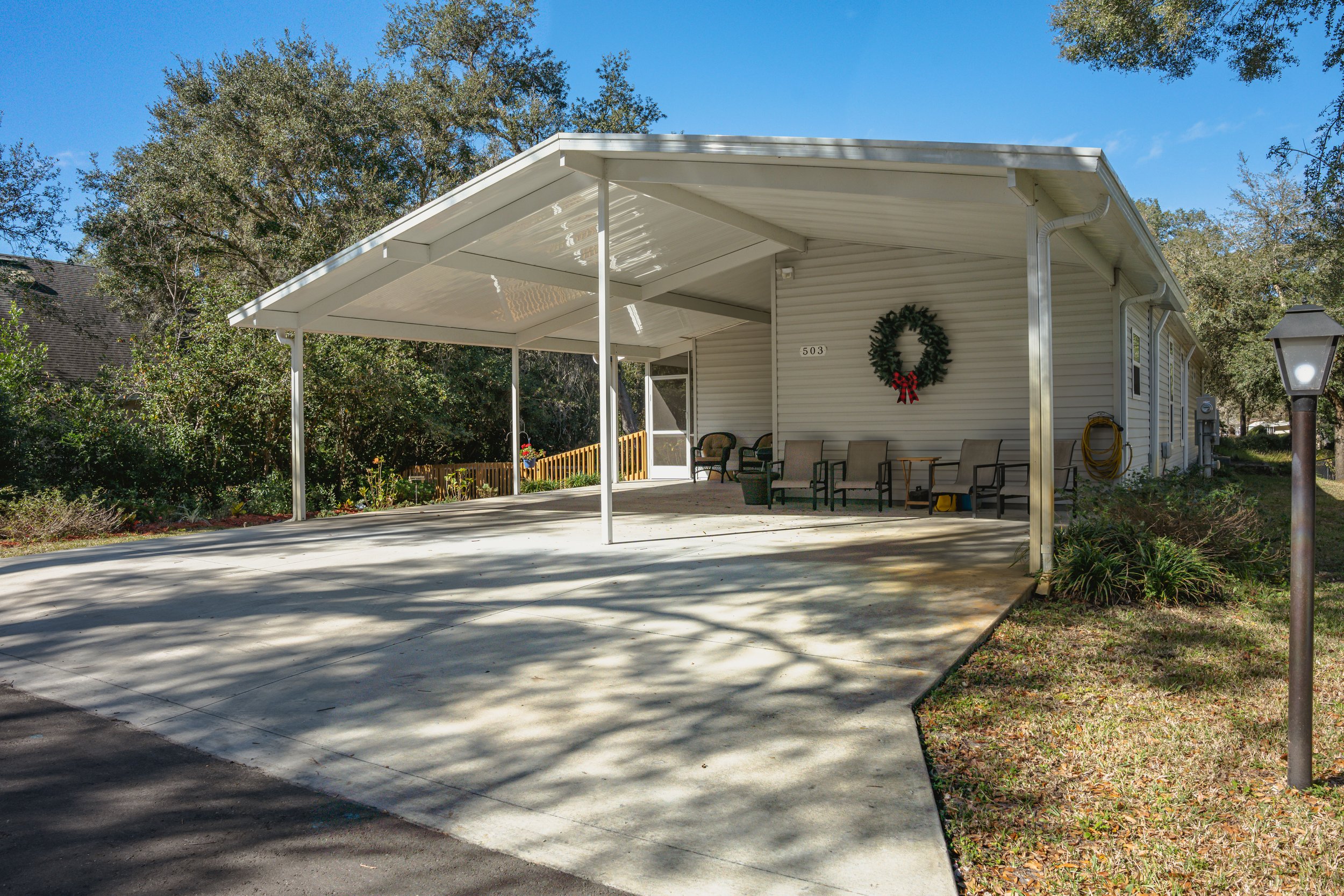 Front of a white house with a porch decorated with a Christmas wreath and outdoor chairs, surrounded by trees and a lawn, under a blue sky.