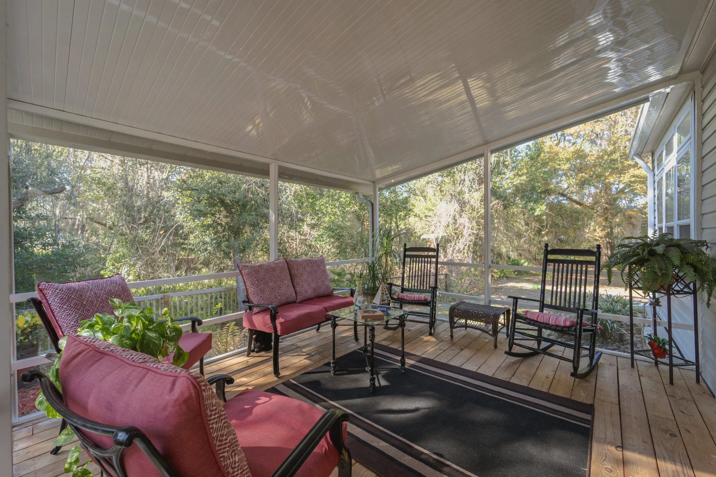  screened porch with pink cushioned seating, black rocking chairs, wicker stool, potted plants, and outdoor scenery with trees