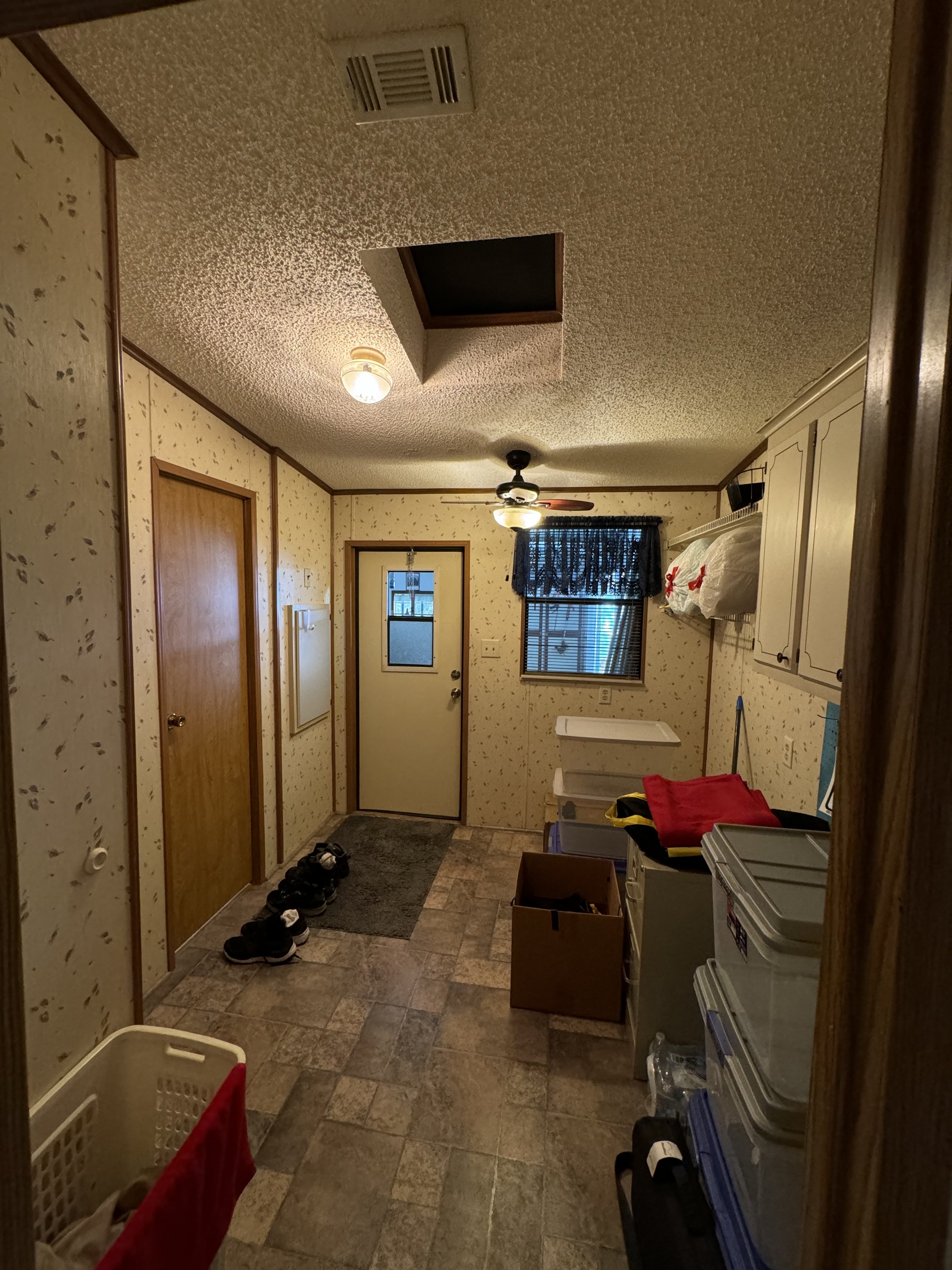 The image shows an entryway with a tiled floor, a door with a window, a window with a curtain, ceiling light, ceiling fan, storage cabinets, cardboard box, plastic storage bins, and shoes on a mat near the door.