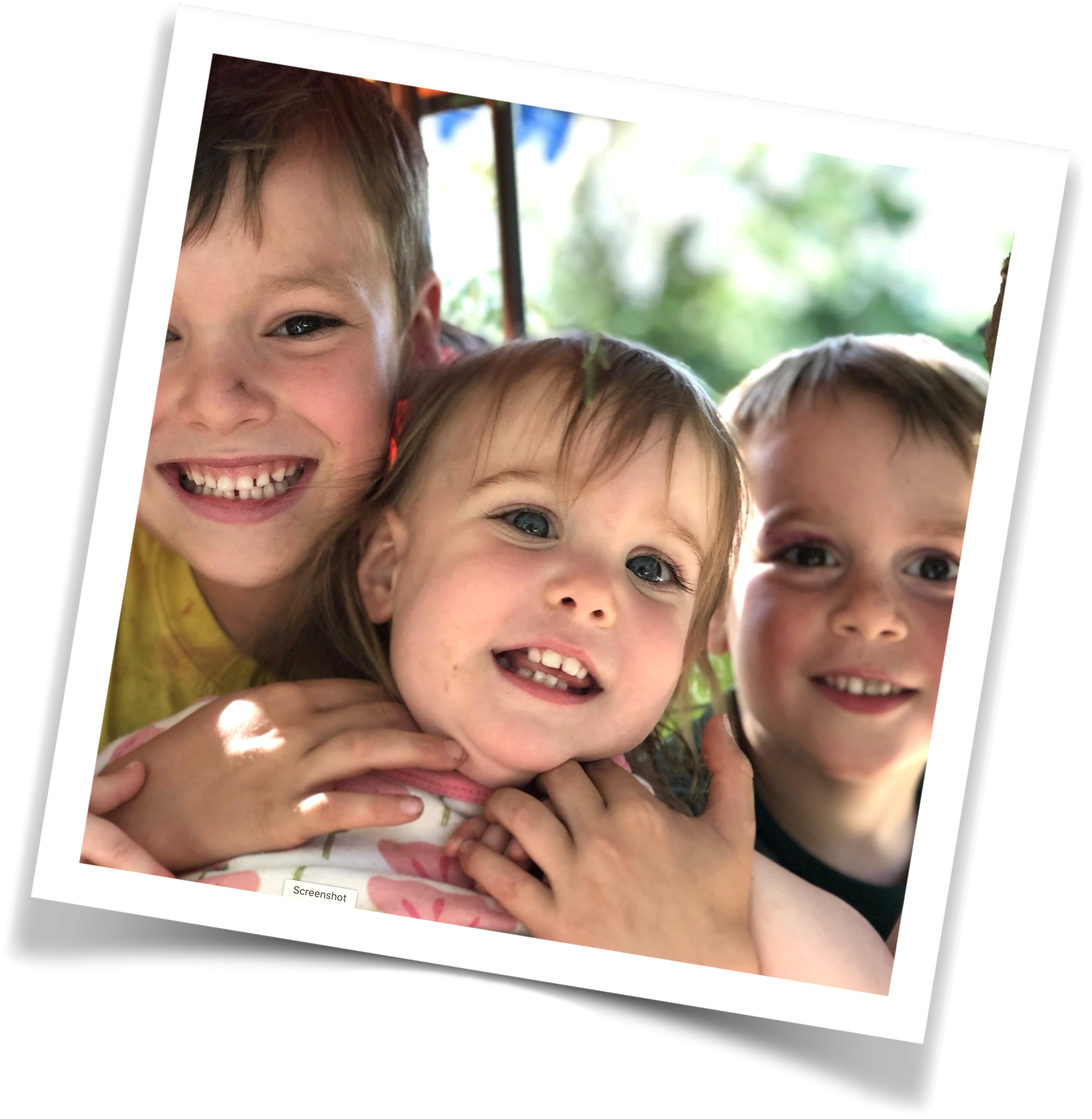 Close-up of three smiling children outside, with a green background and sunlight.