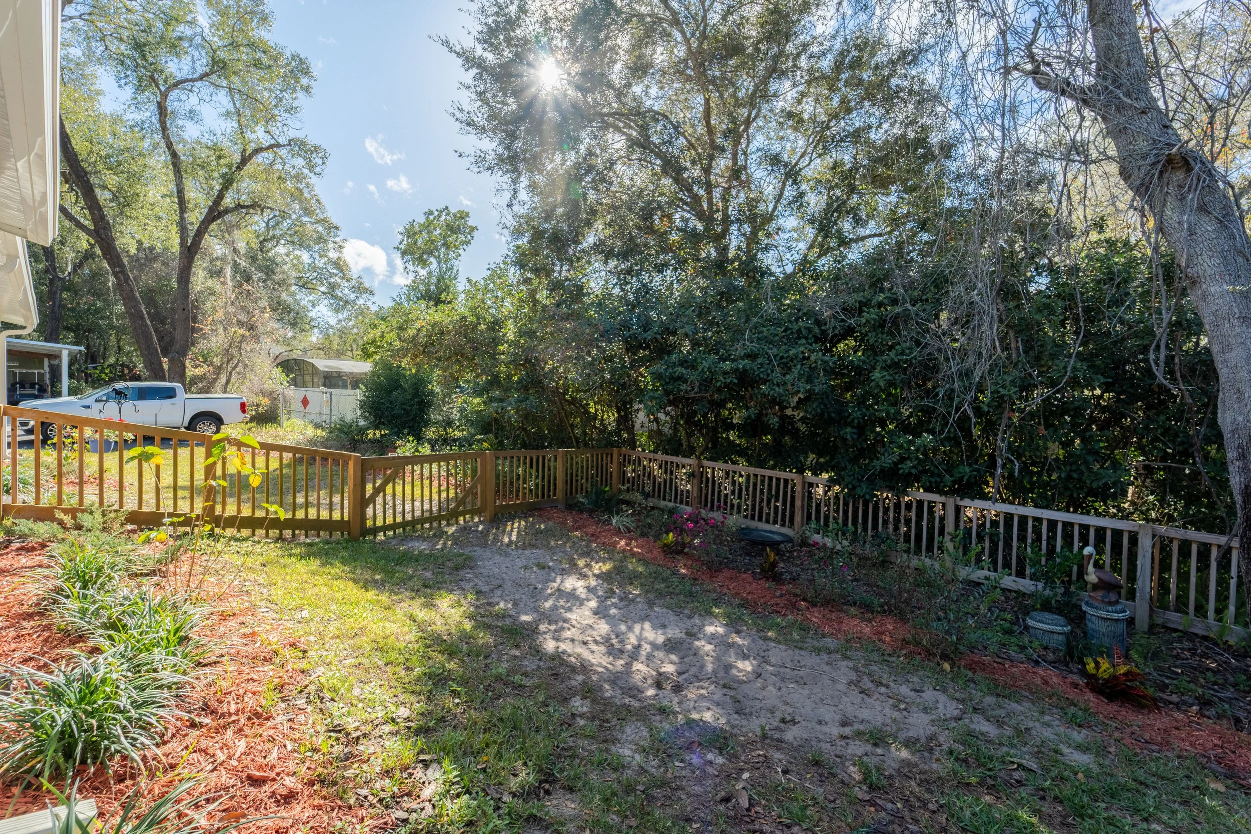 Backyard with a wooden fence, trees, a parked white truck, and backyard decorations under a bright sun.